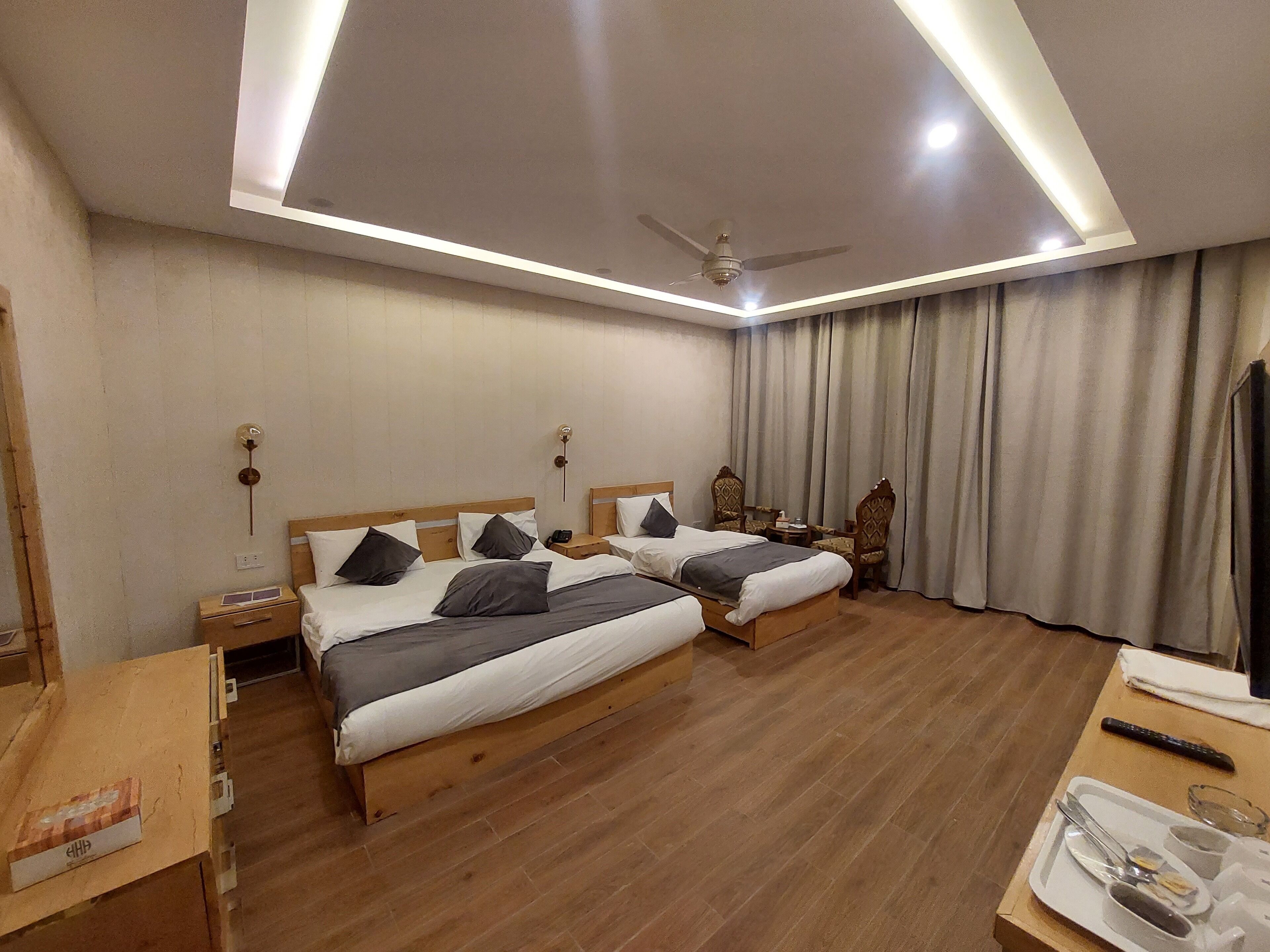 Habitación de hotel con dos camas de madera, un ventilador de techo, un escritorio con silla y una ventana con cortinas.