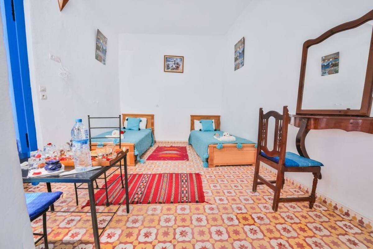 A small, cozy room with two beds, a desk, and a chair. There is a mirror on the wall and a small table with a water bottle and other items.