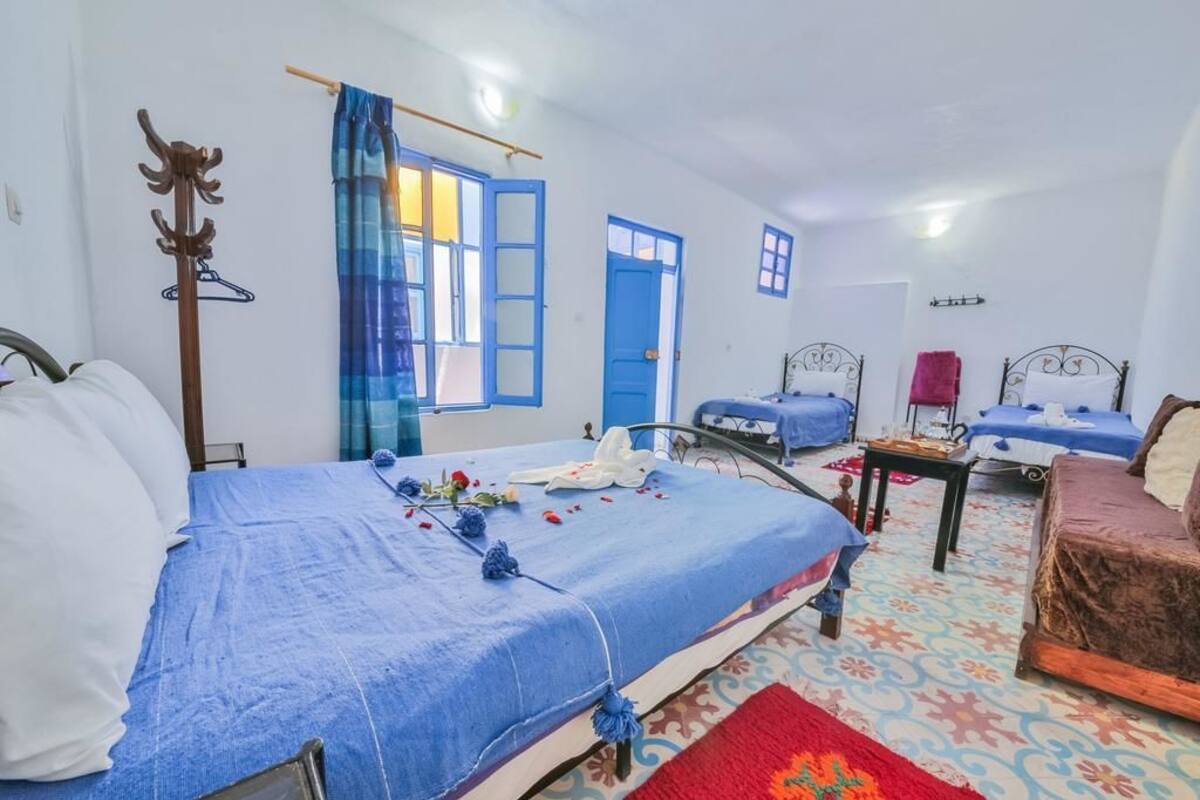 A hotel room with a large bed, two smaller beds, a wooden coat rack, and a window with blue curtains.