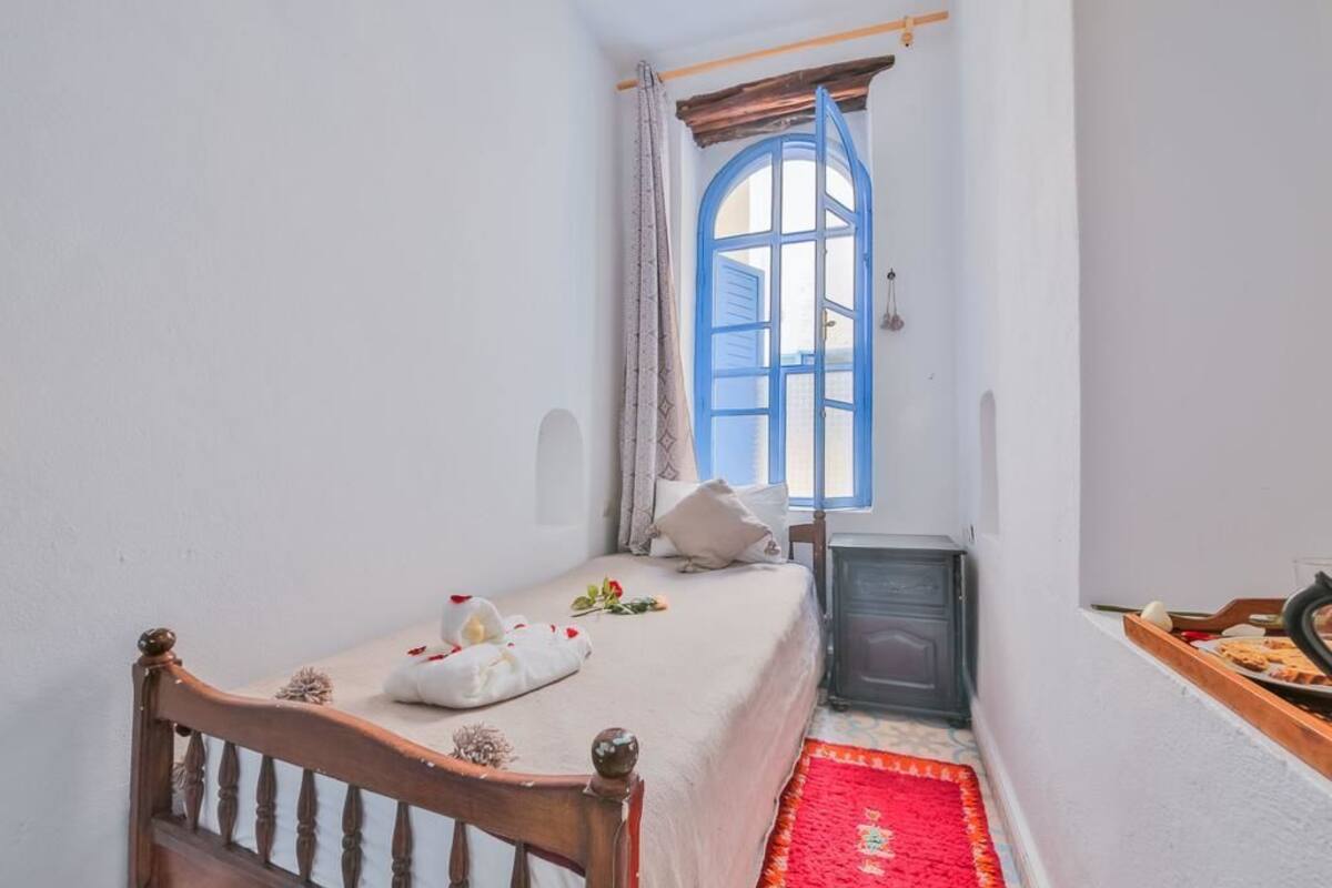 A small, cozy bedroom with a single bed, a wooden headboard, a red rug, a small table with food, and a window with blue shutters.