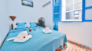 A bedroom with a bed, blue bedding, a window with blue shutters, a wall-mounted air conditioner, and a framed picture. - Riad Aylal (Essaouira)