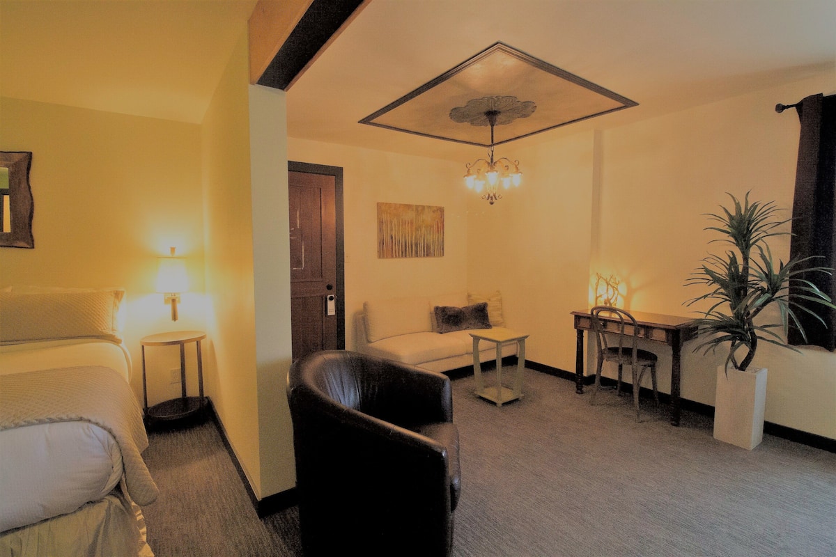 A hotel room with a bed, a chair, a small table, a desk, a potted plant, and a ceiling light fixture.