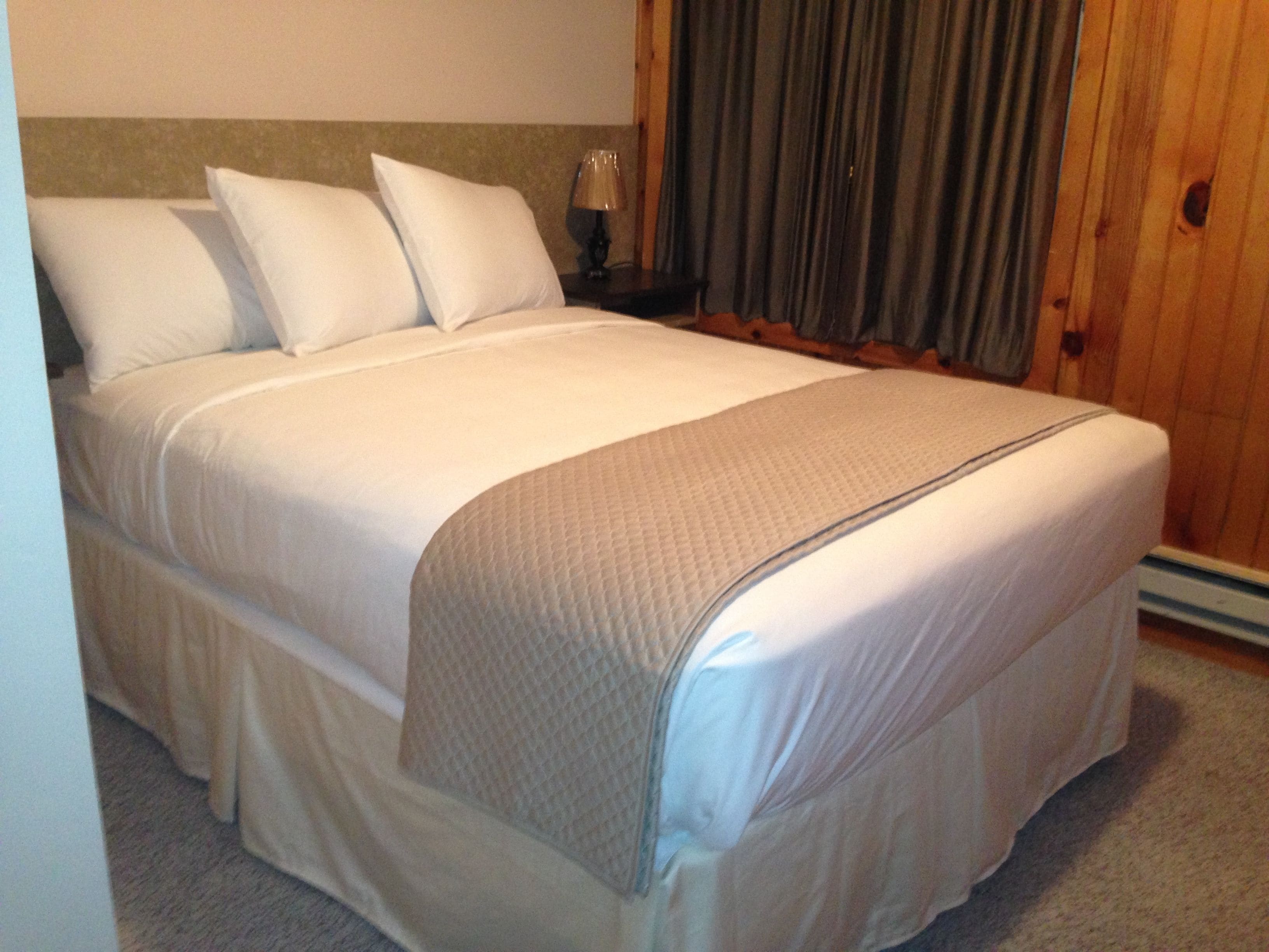 A neatly made bed with white linens and a beige throw blanket, a wooden headboard, a nightstand with a lamp, and a window with curtains.