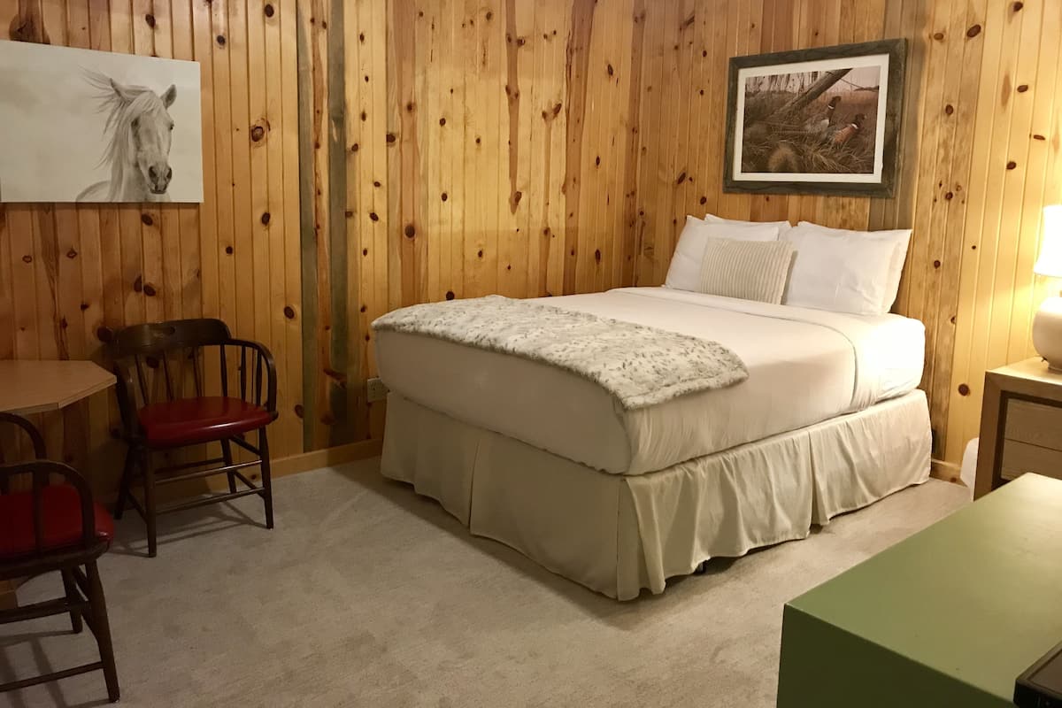 A room with a wooden paneled wall, a bed with white bedding, a chair, a small table, and a framed picture on the wall.