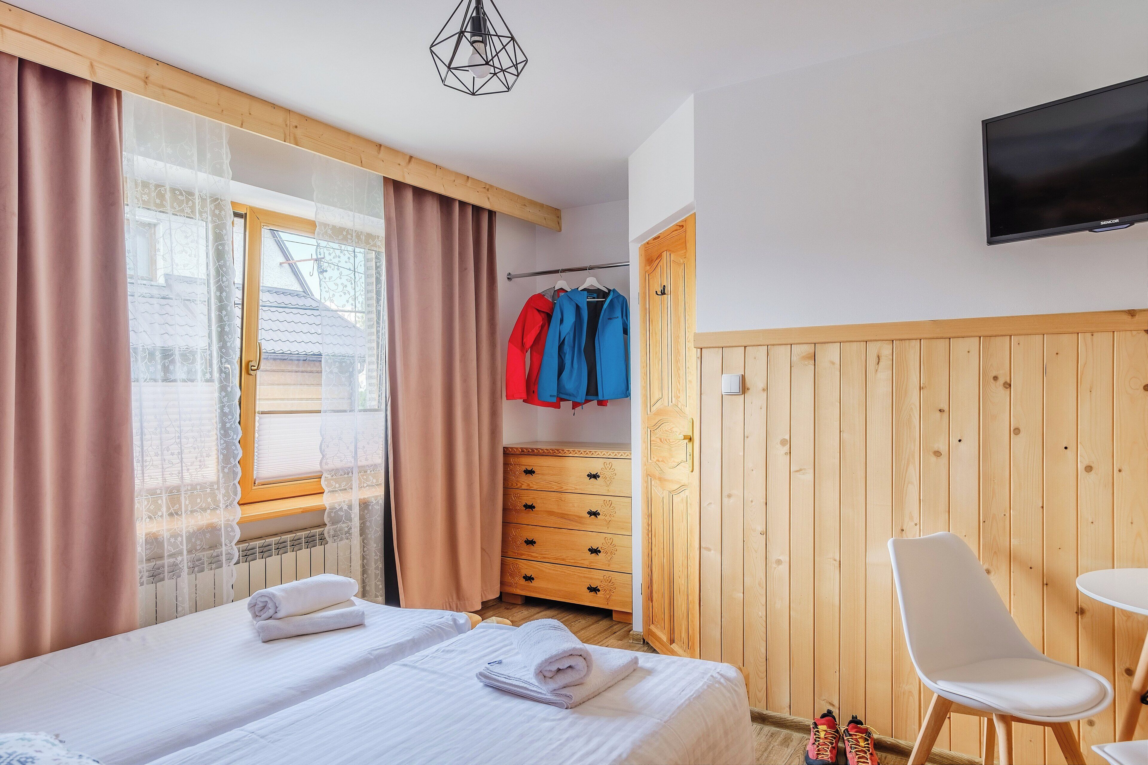 A hotel room with a bed, a wooden wardrobe, a television mounted on the wall, and a window with curtains.