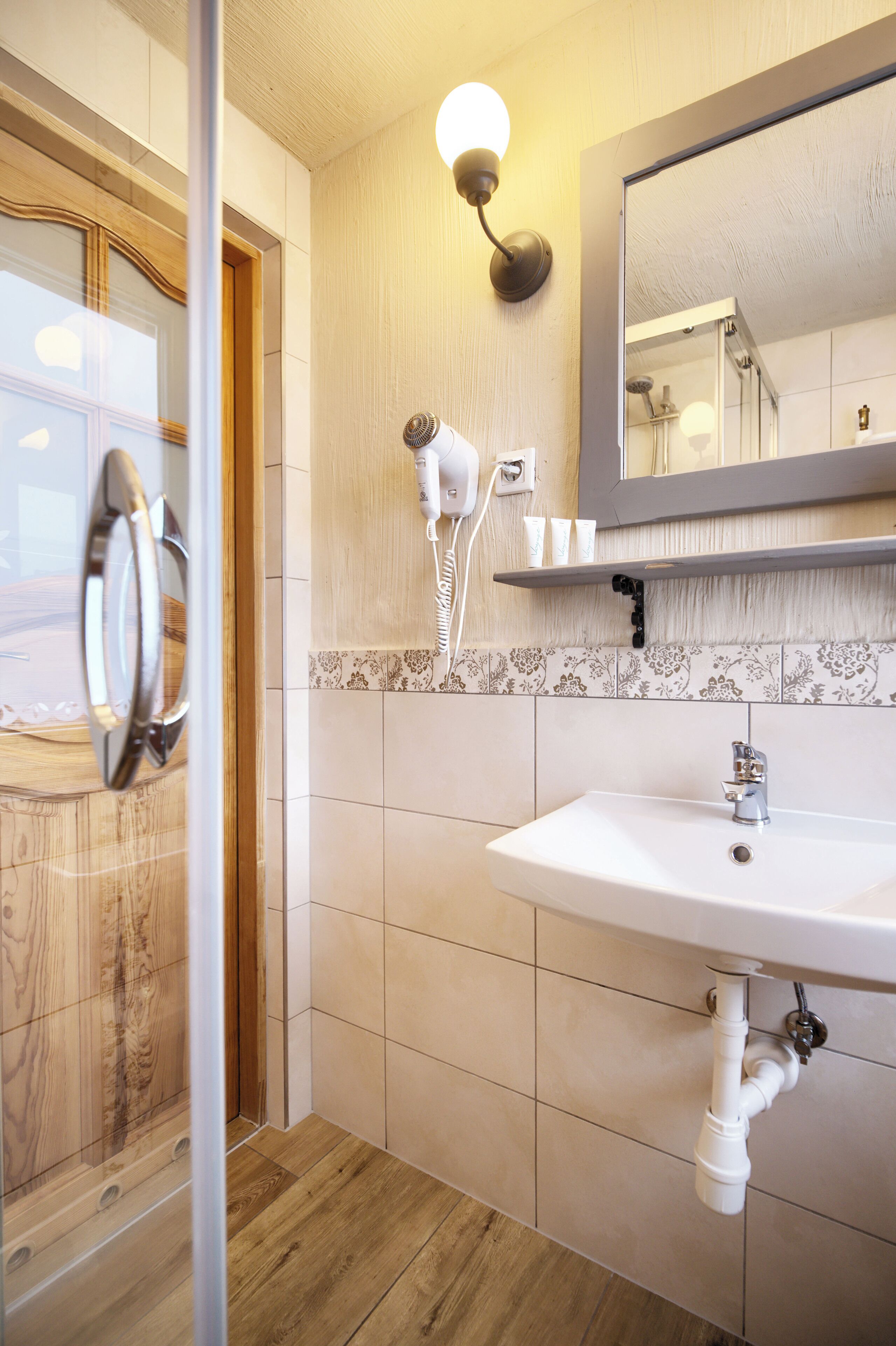 Un baño con suelo de madera, un lavamanos blanco, un espejo y un secador de pelo fijado a la pared.