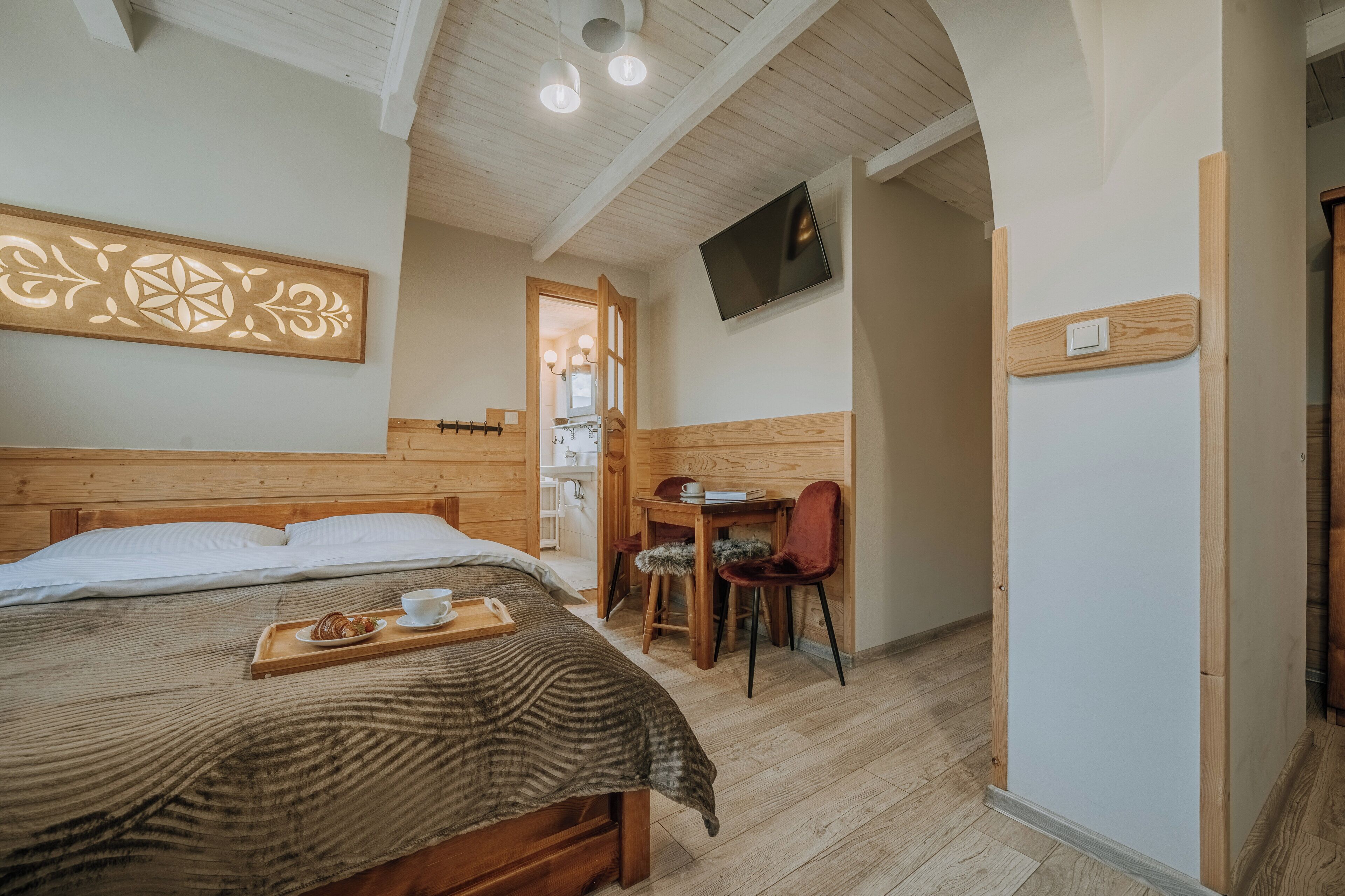 A wooden-framed room with a bed, a desk, a chair, a TV mounted on the wall, and a door leading to another room.