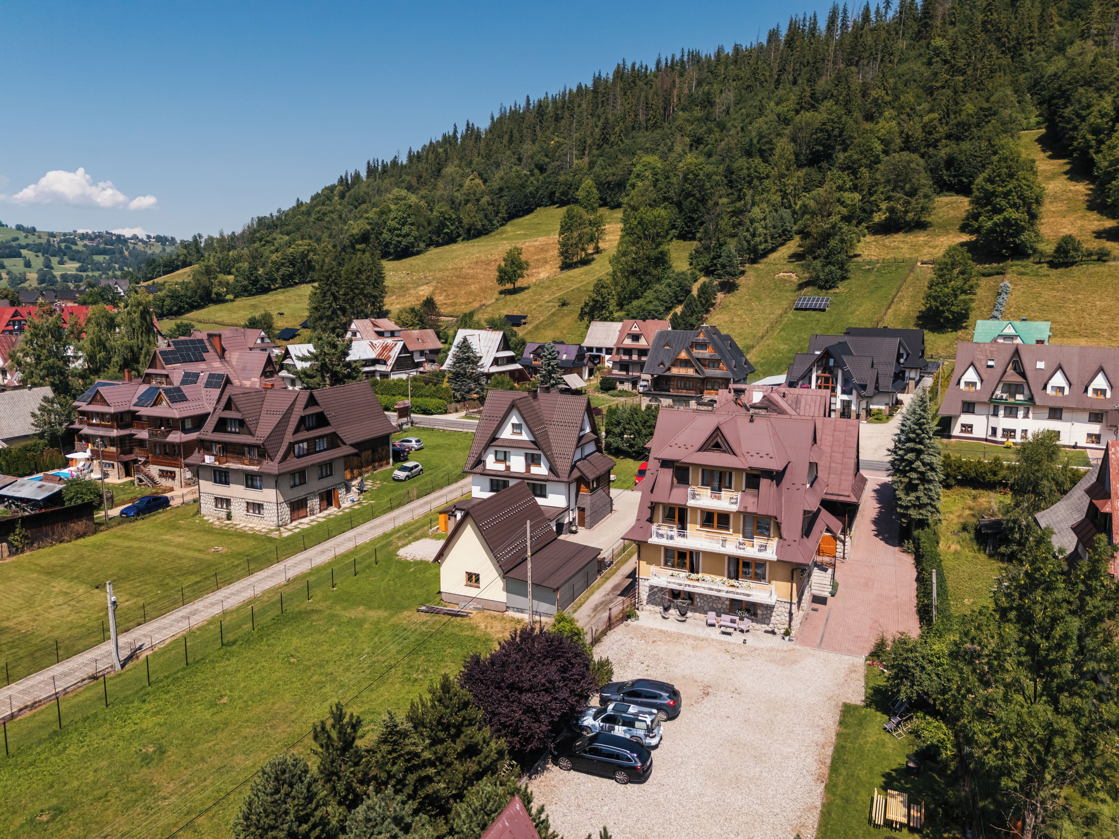 A residential area with houses featuring sloped roofs and solar panels, surrounded by greenery and hills.