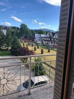 A view from a balcony overlooking a village with traditional houses, a car parked in front, and a mountain range in the background.