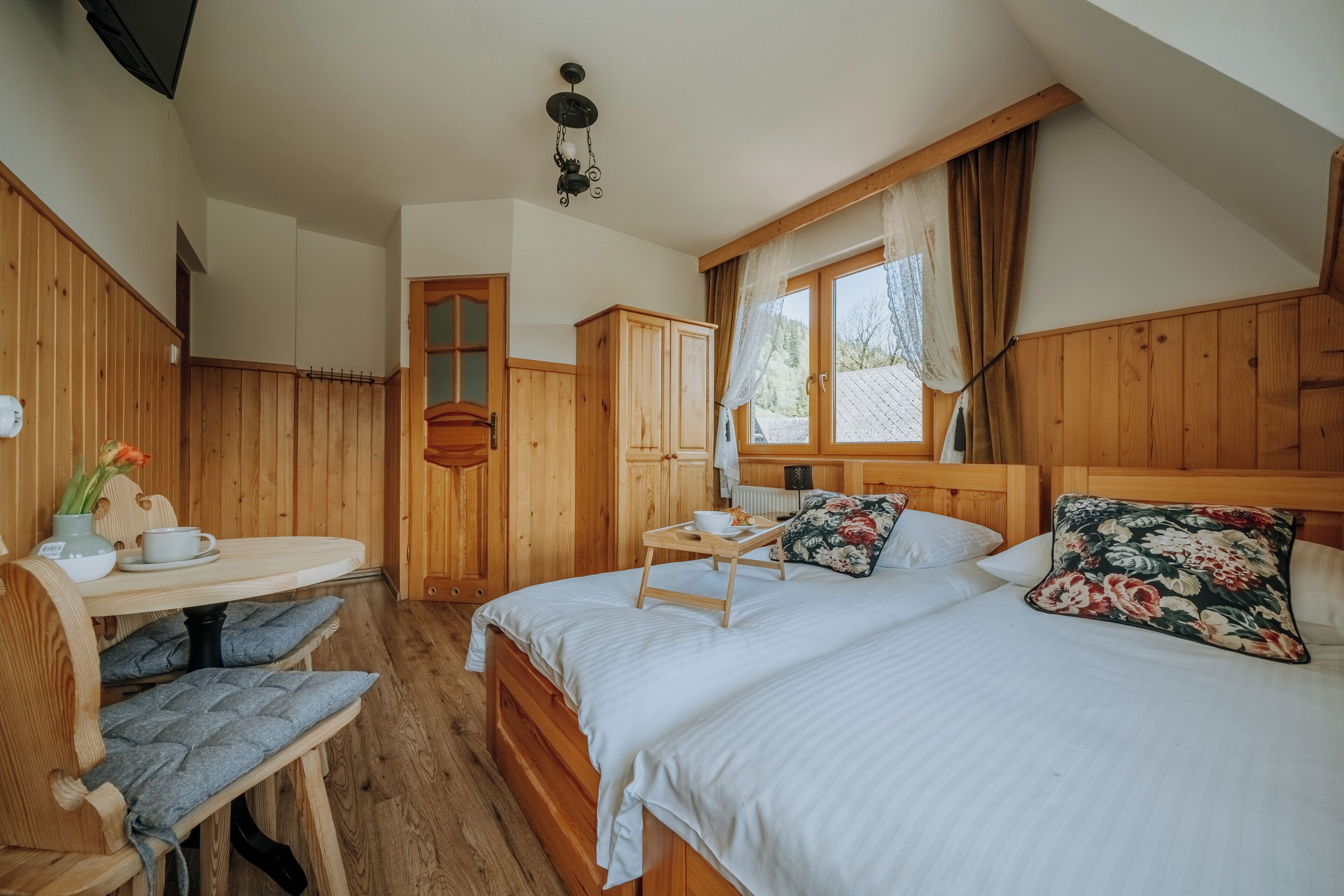 A wooden cabin bedroom with a bed, a small table with a cup and saucer, a chair, and a window with curtains.