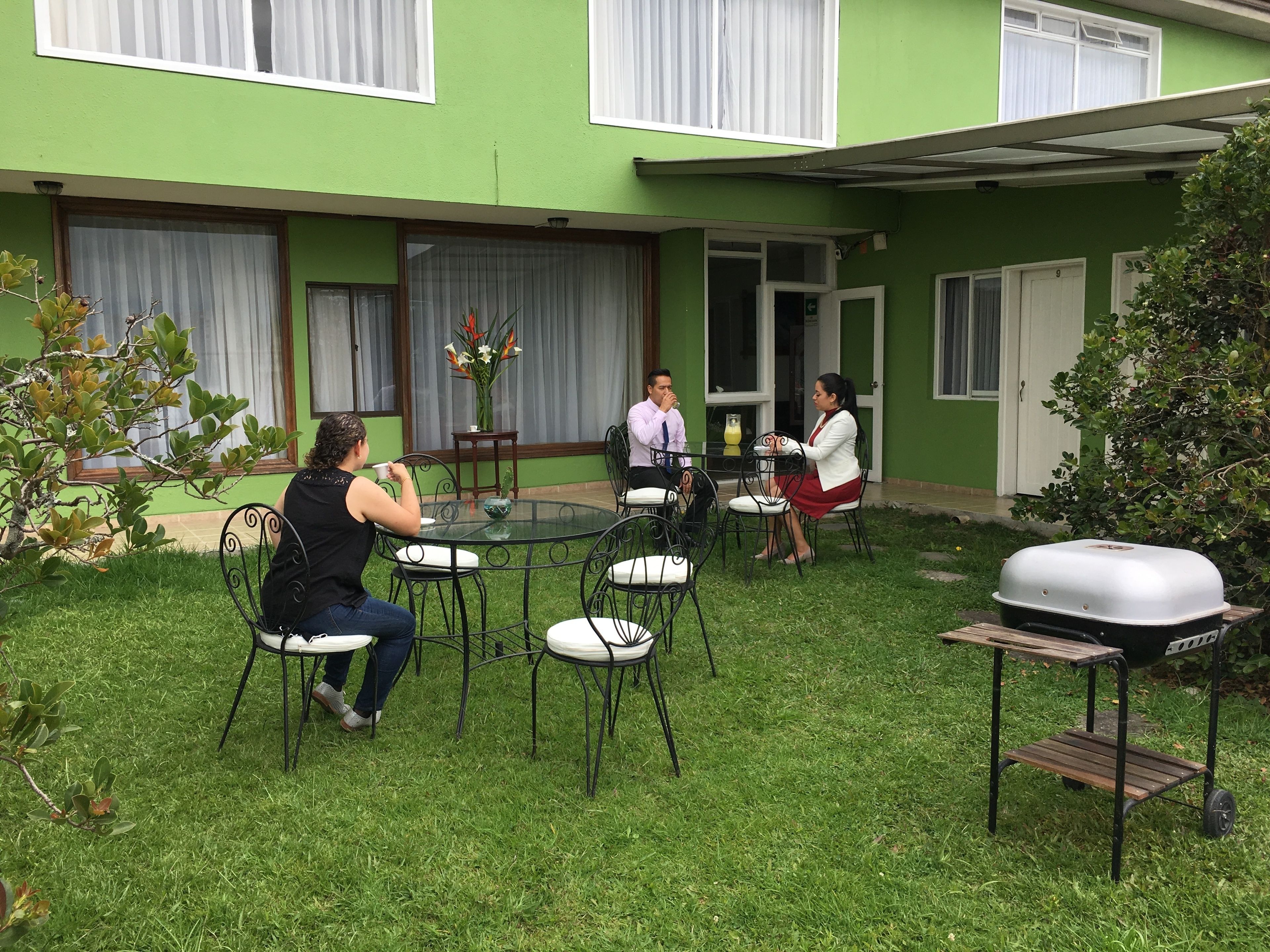 Een terras met een ronde tafel en stoelen, een barbecue, en een kleine tuin met een boom en een struik.