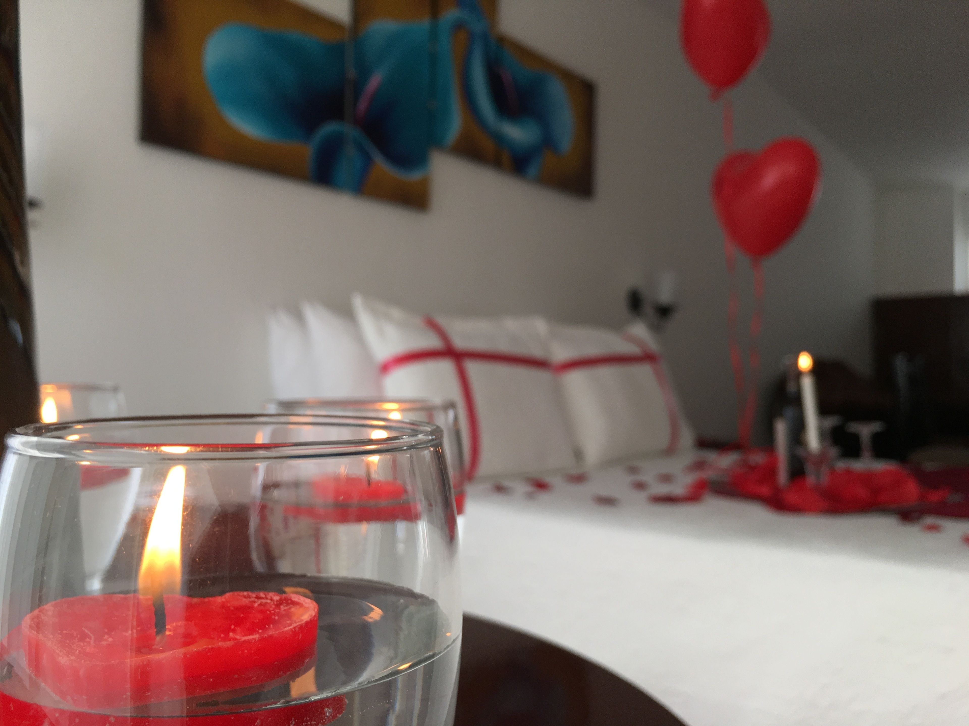 A candle in a glass holder, a bed with white linens and red heart decorations, and a painting on the wall.