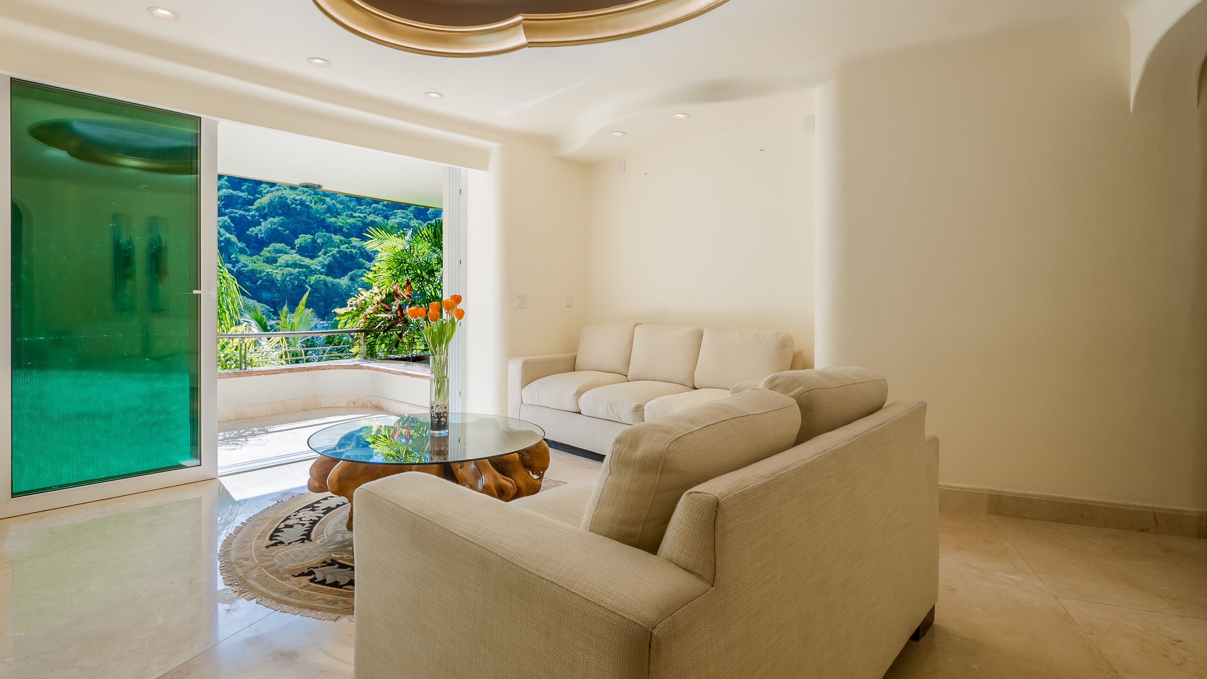 Un salon moderne avec une table en verre, un canapé beige et une vue sur une végétation luxuriante.