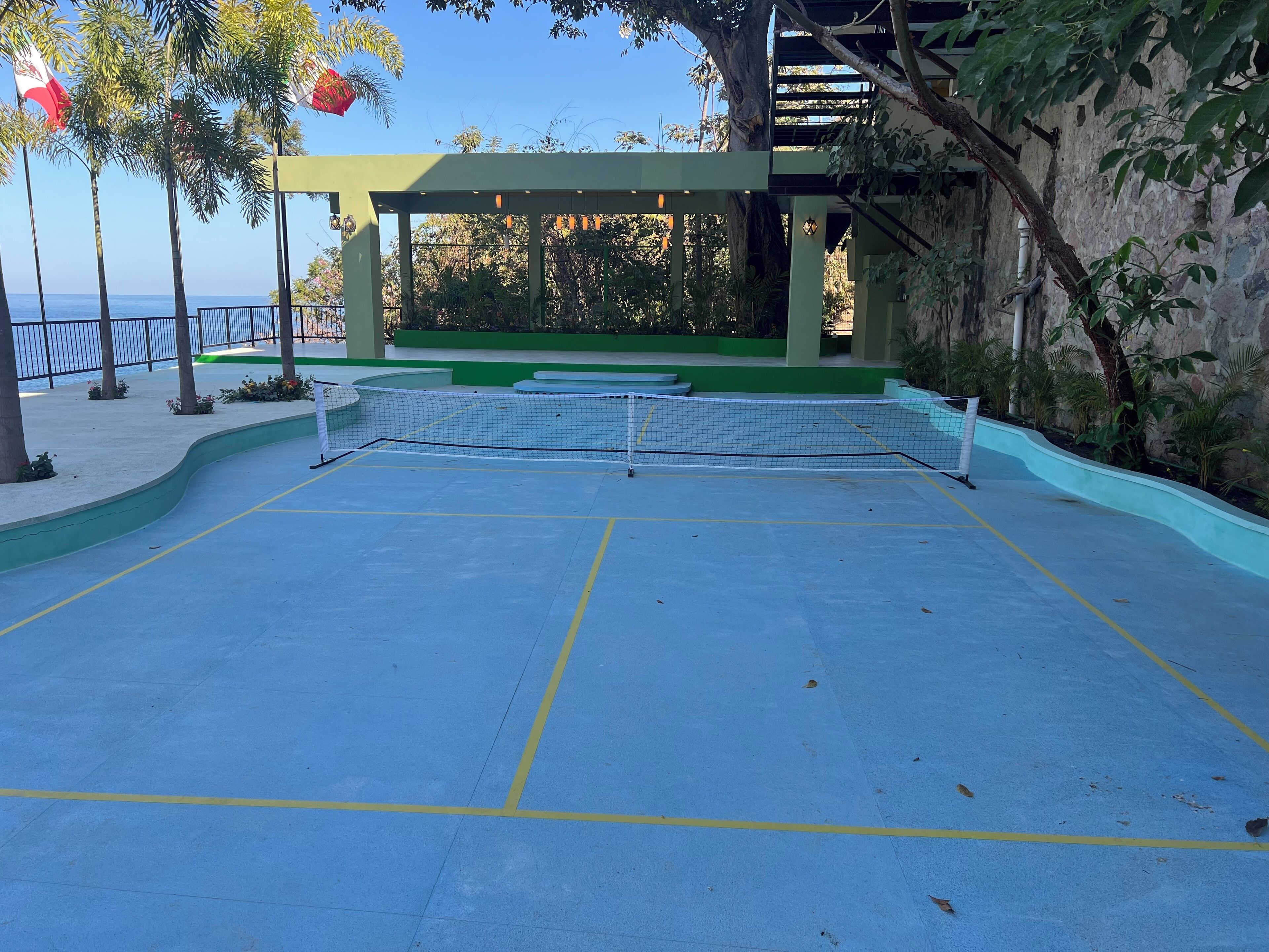 Una cancha de tenis al aire libre con una red, rodeada de vegetación y un muro de piedra.