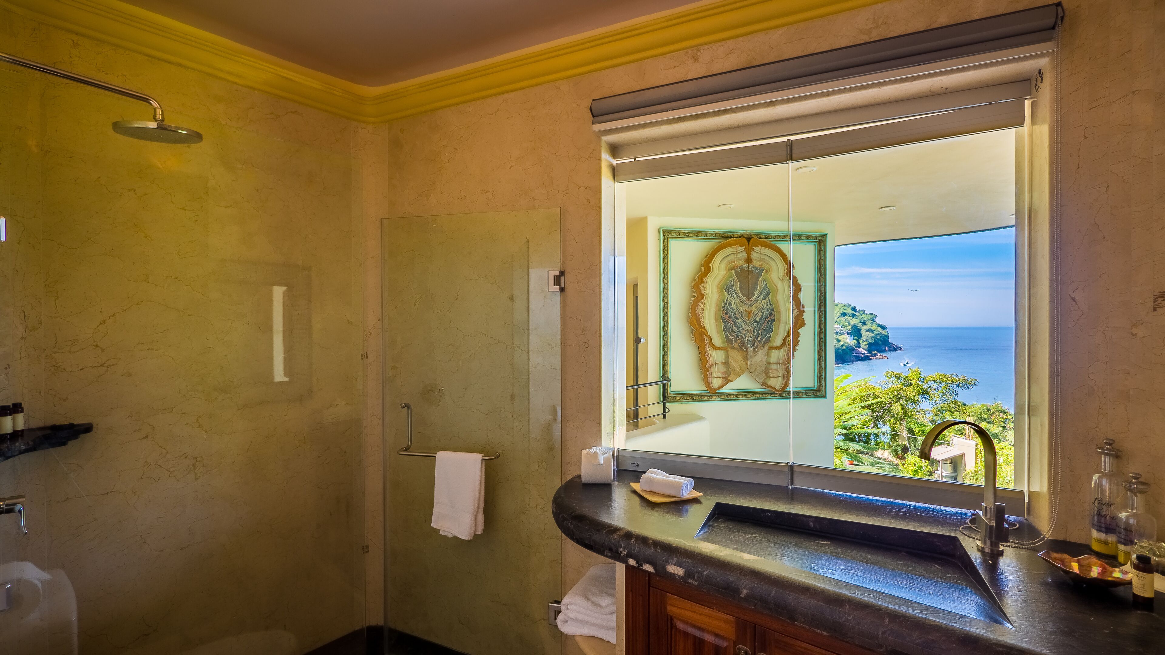 A bathroom with a shower, a sink with toiletries, and a large mirror reflecting a view of the sea.