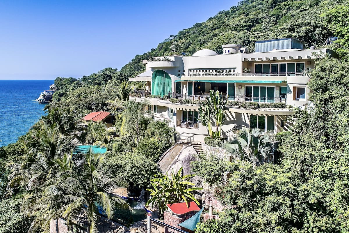 Un resort de varios pisos con piscina, rodeado de exuberante vegetación y vistas al mar.