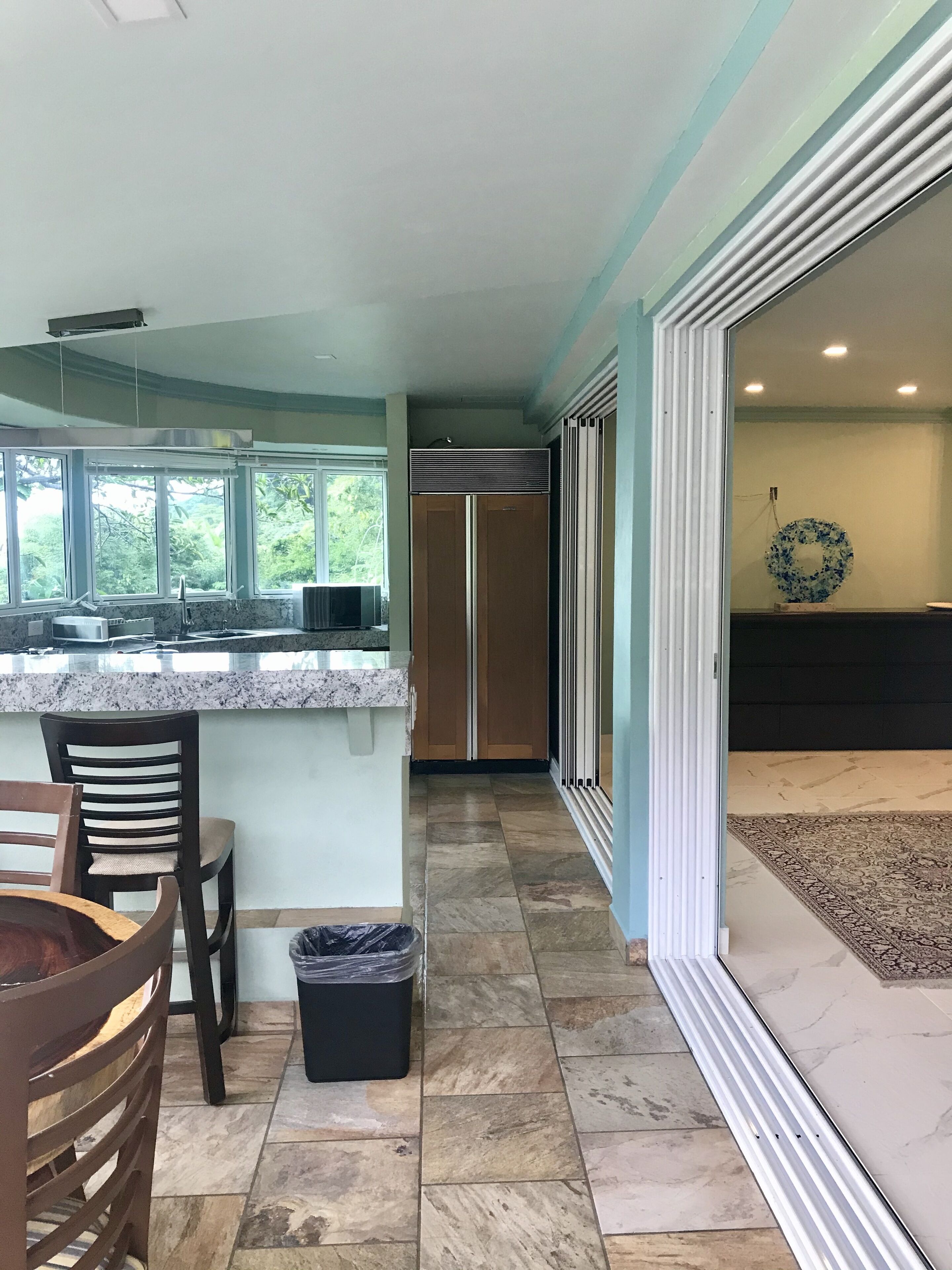A kitchen with a marble countertop, wooden chairs, a trash bin, and a view of a dining area with a rug and a wall decoration.