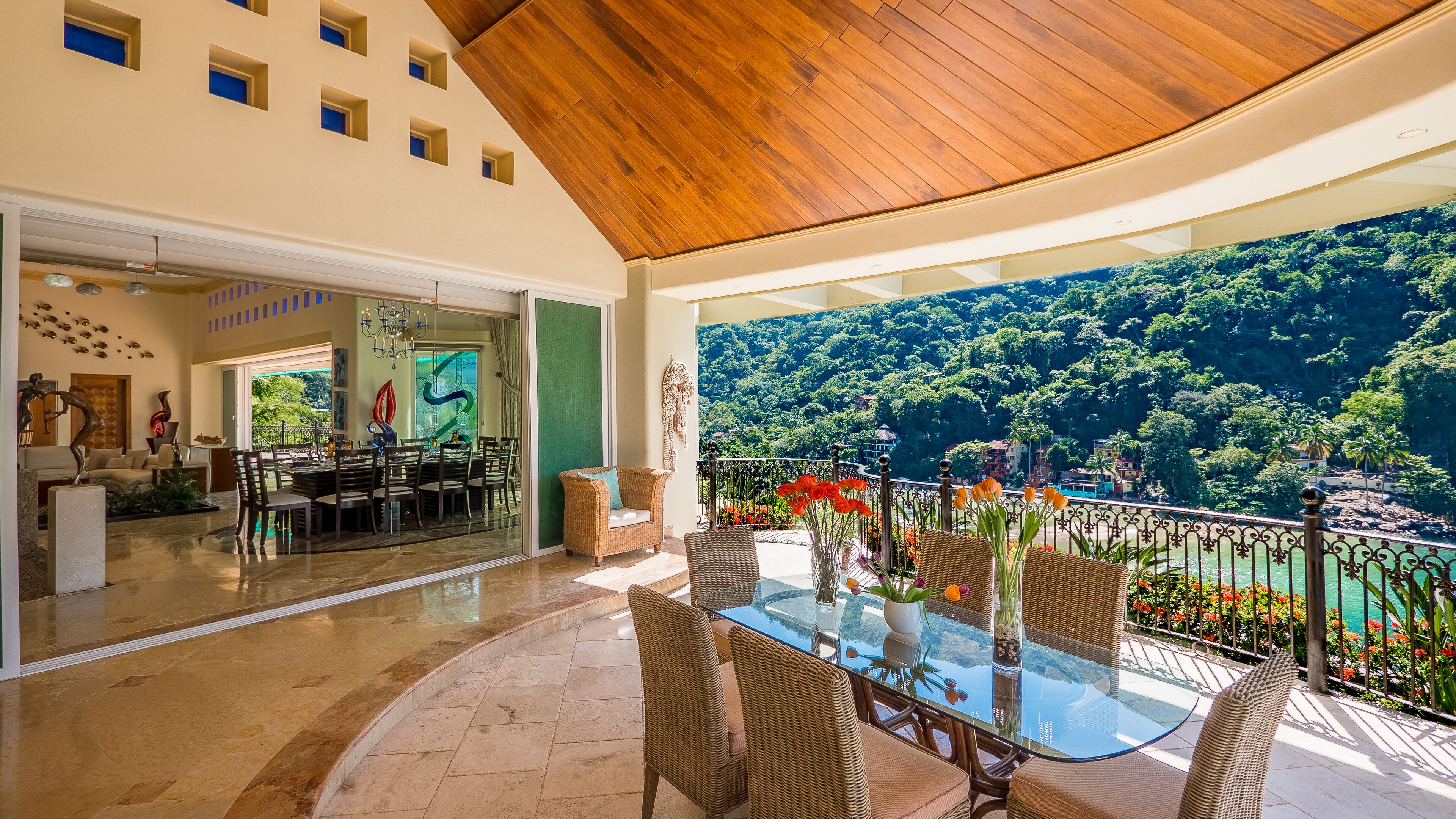 Un espacio interior amplio con una mesa y sillas de comedor, un ventanal grande que ofrece una vista panorámica de un bosque exuberante y un río, y un techo con diseño de paneles de madera.
