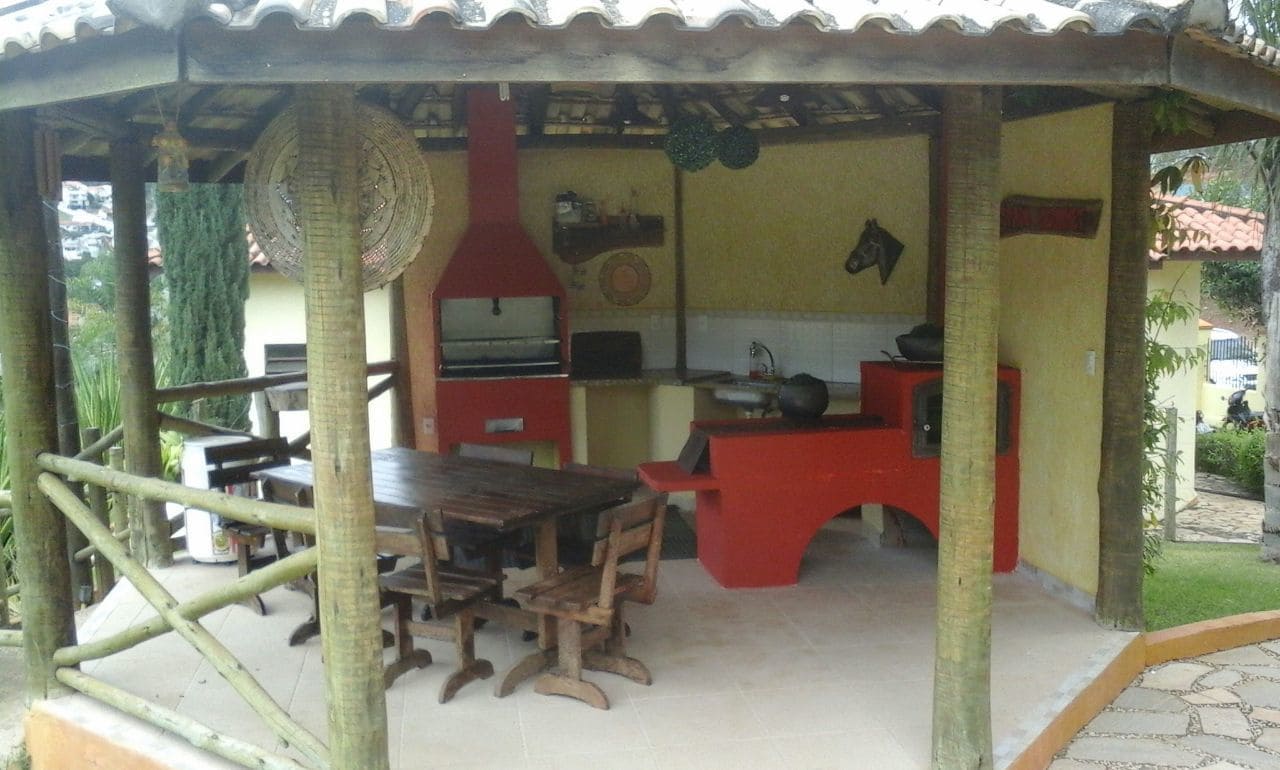 Uma área de refeições ao ar livre coberta, com mesa e cadeiras de madeira, churrasqueira vermelha e piso de azulejos.
