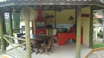 Uma área de refeições ao ar livre coberta, com mesa e cadeiras de madeira, churrasqueira vermelha e piso de azulejos.
