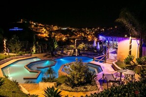 Vista nocturna d'un complex turístic amb una gran piscina de múltiples arcs, envoltada de mobiliari exterior i palmeres.