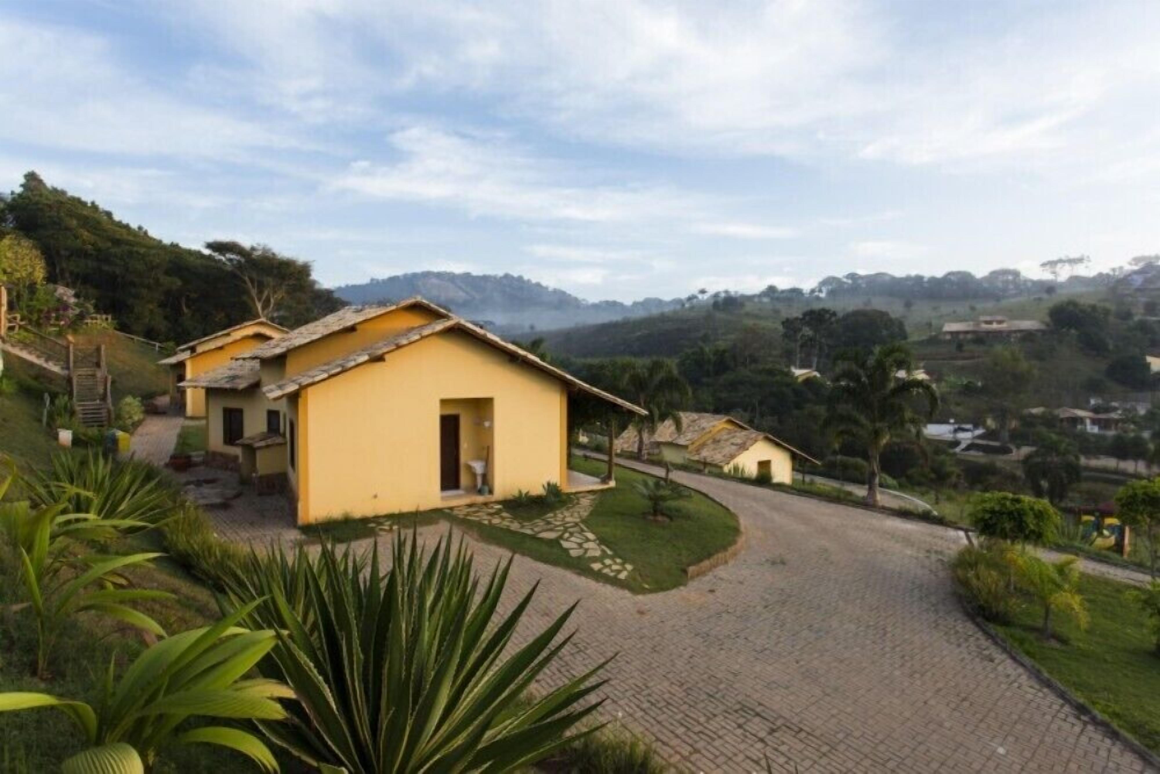 A yellow building with a curved driveway, surrounded by greenery and hills in the background.