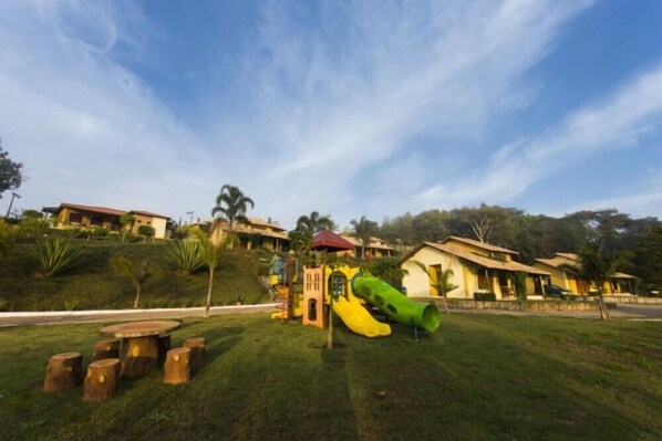 A playground with slides and climbing structures, surrounded by grass and trees, with houses in the background.