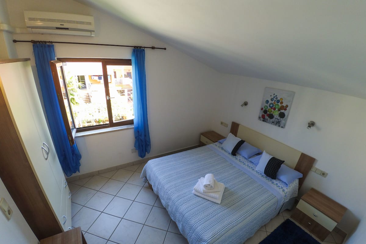 A bedroom with a bed, a nightstand, a window with curtains, a wall-mounted air conditioner, and a small rug.
