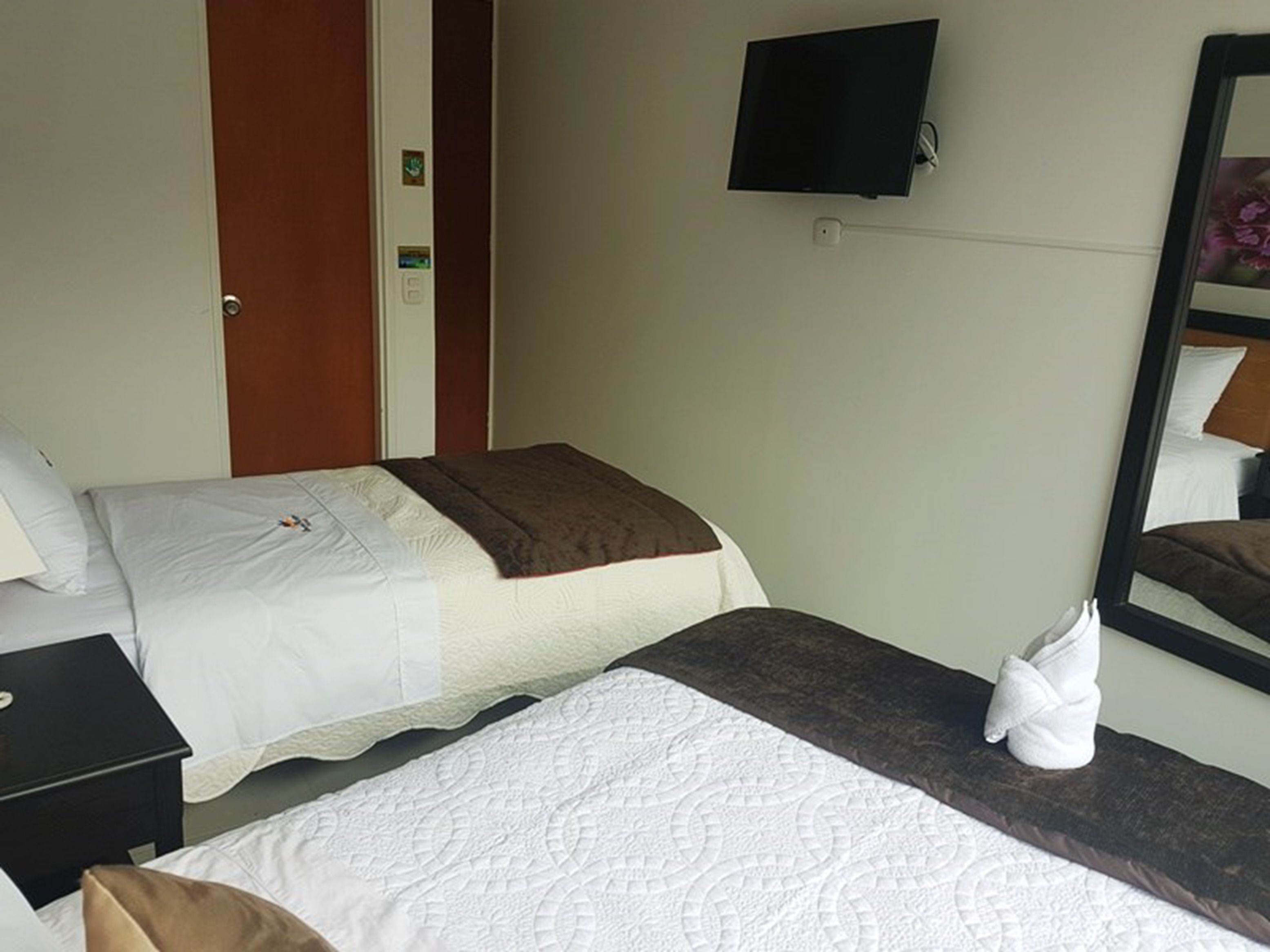 Habitación de hotel con dos camas, un televisor montado en la pared, un espejo y una toalla sobre la cama.