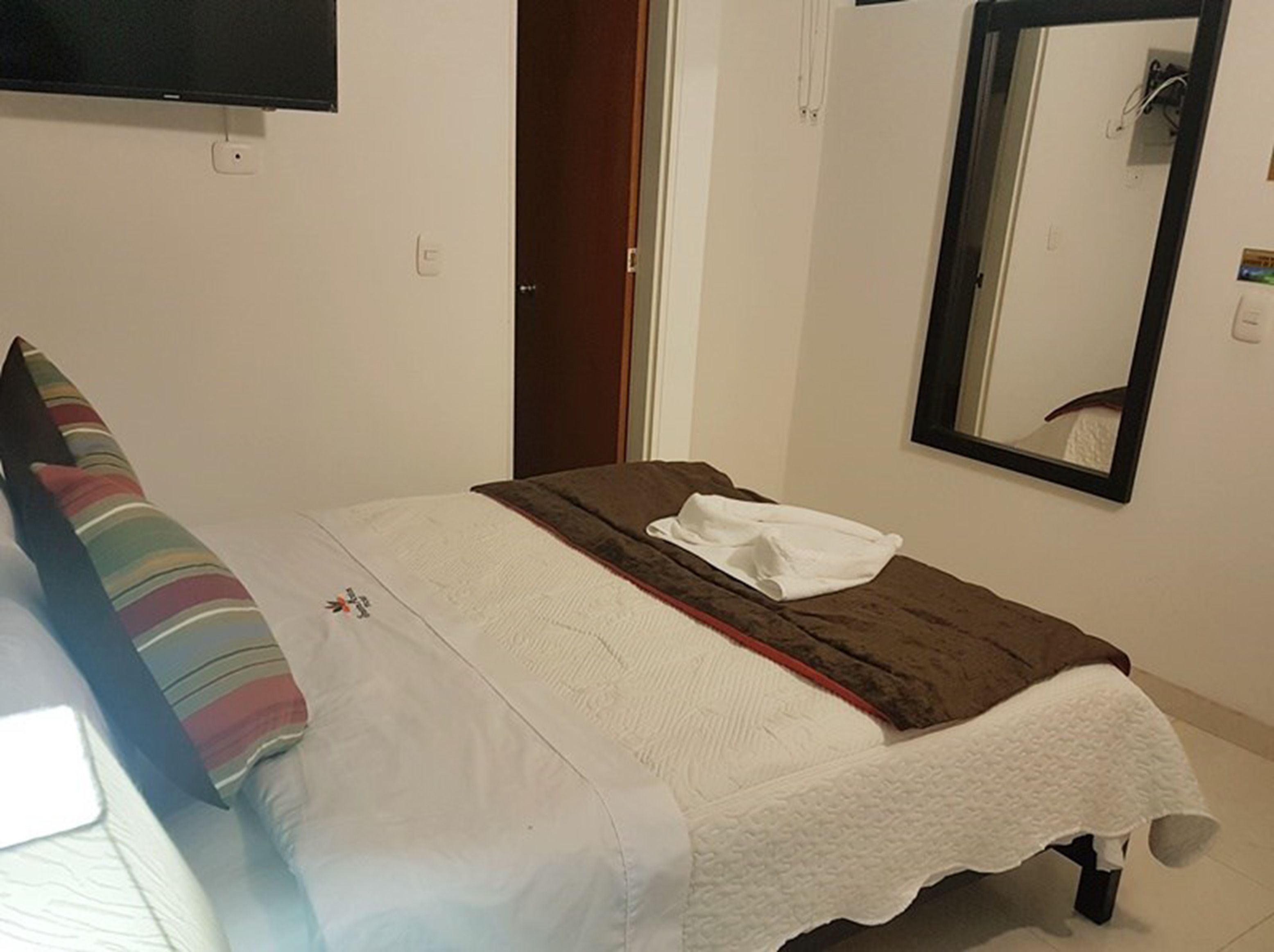 Una cama individual con una colcha blanca y una almohada a rayas, una manta marrón y una toalla blanca sobre la cama. Hay un televisor montado en la pared encima de la cama, una puerta a la derecha y un espejo en la pared al lado de la puerta.