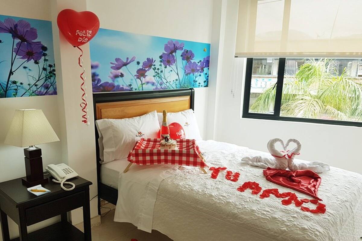 A hotel room with a bed, a nightstand with a lamp and phone, a red heart-shaped balloon, and a painting of flowers.