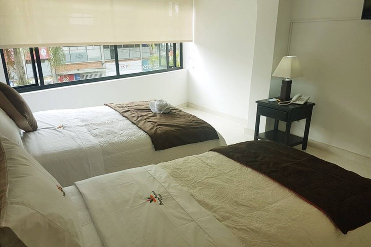 A hotel room with two beds, a nightstand with a lamp, and a window with a view of the outside.