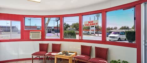 Una habitación con cuatro sillas tapizadas rojas, una mesa de madera y ventanas grandes con un letrero de 'NO VACANCY'.