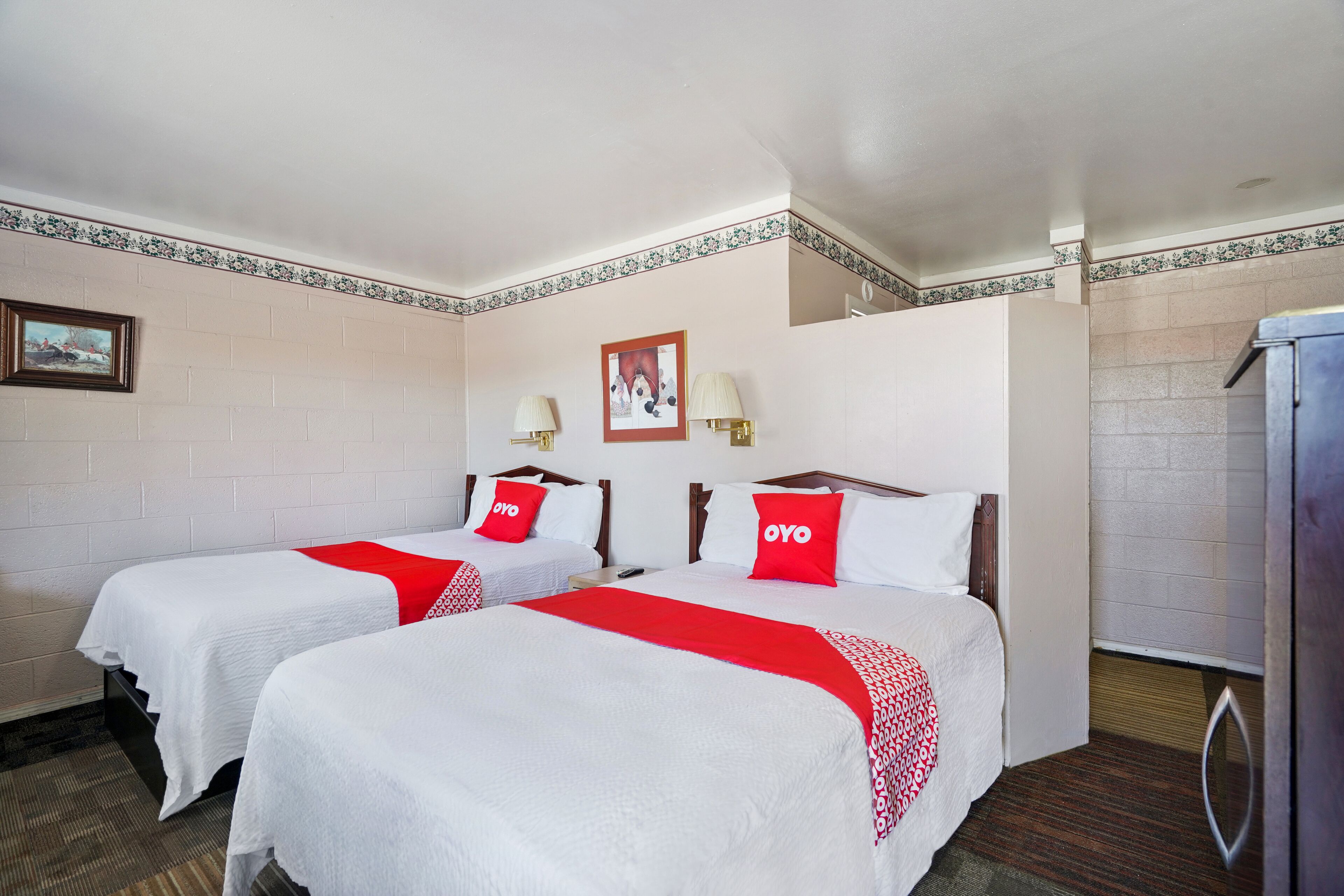 Een hotelkamer met twee bedden, elk met witte beddengoed en rode accenten, een klein schilderij aan de muur en een koelkast in de hoek.