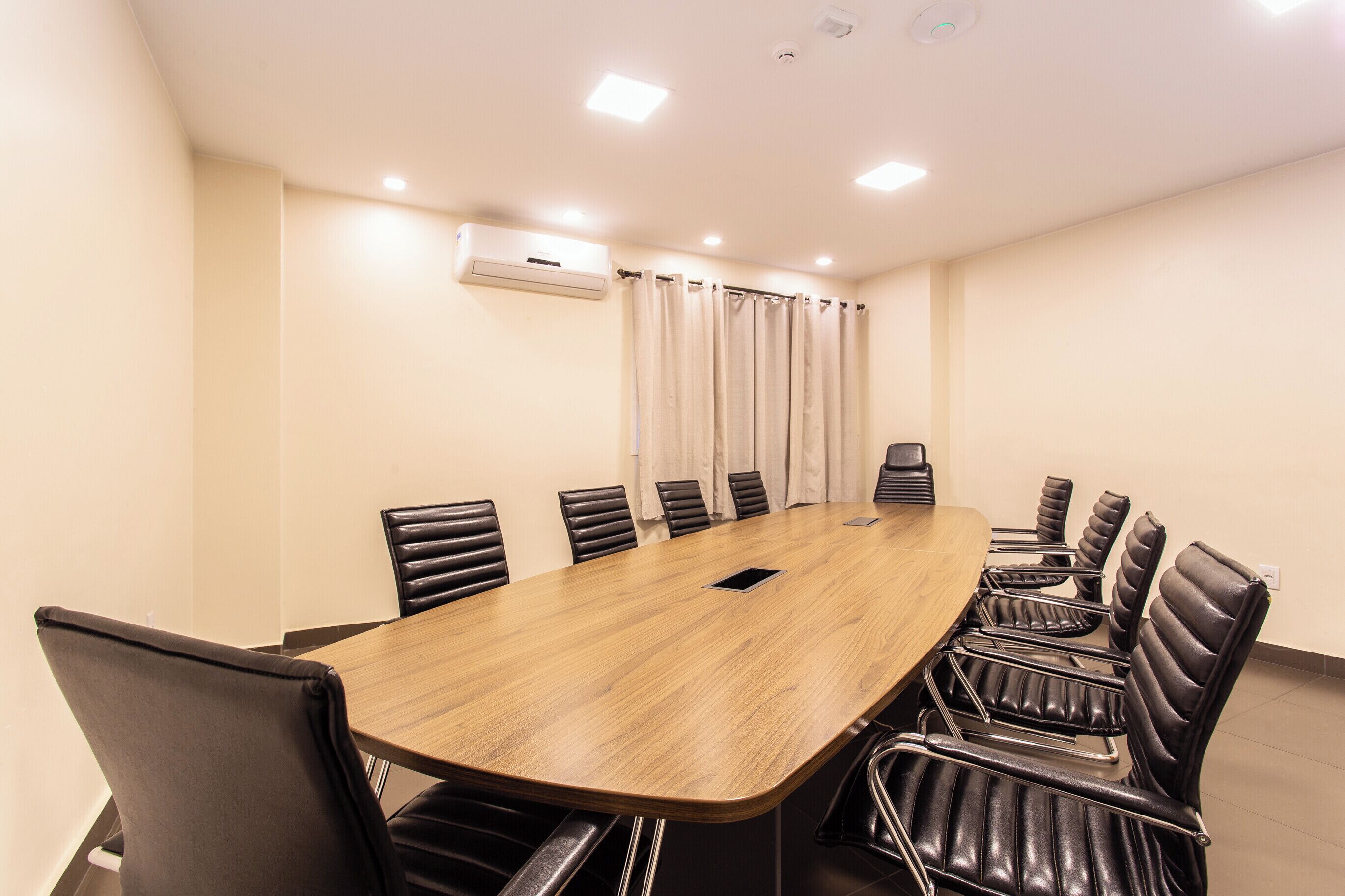 A conference room with a long wooden table, black leather chairs, and a white air conditioning unit on the wall.