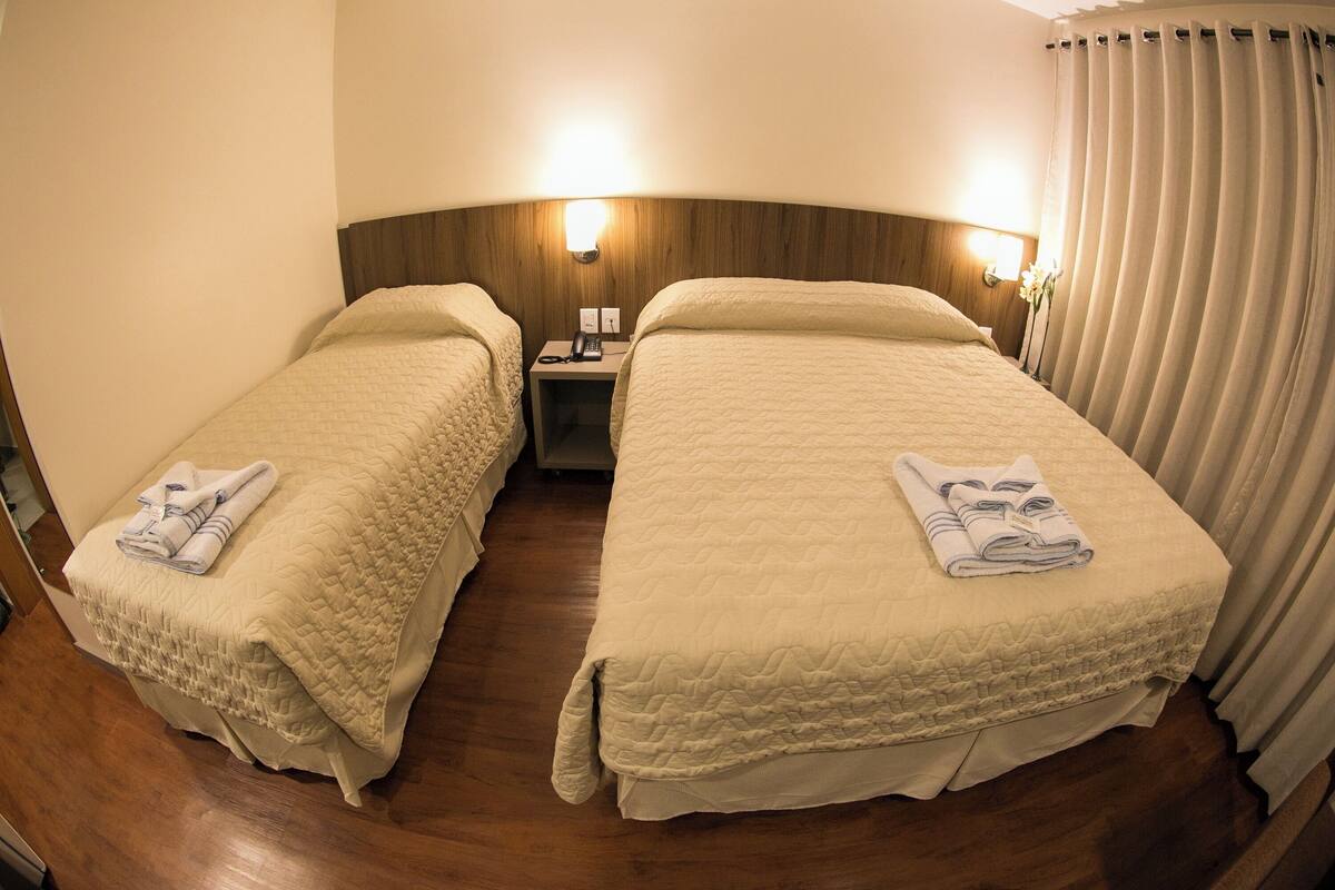 A hotel room with two beds, each with a quilted bedspread and a folded towel on the bed. There is a nightstand with a lamp between the beds, and a curtain on the right side.
