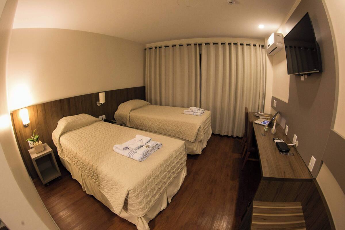 A hotel room with two beds, a desk, a TV, and a window with curtains.