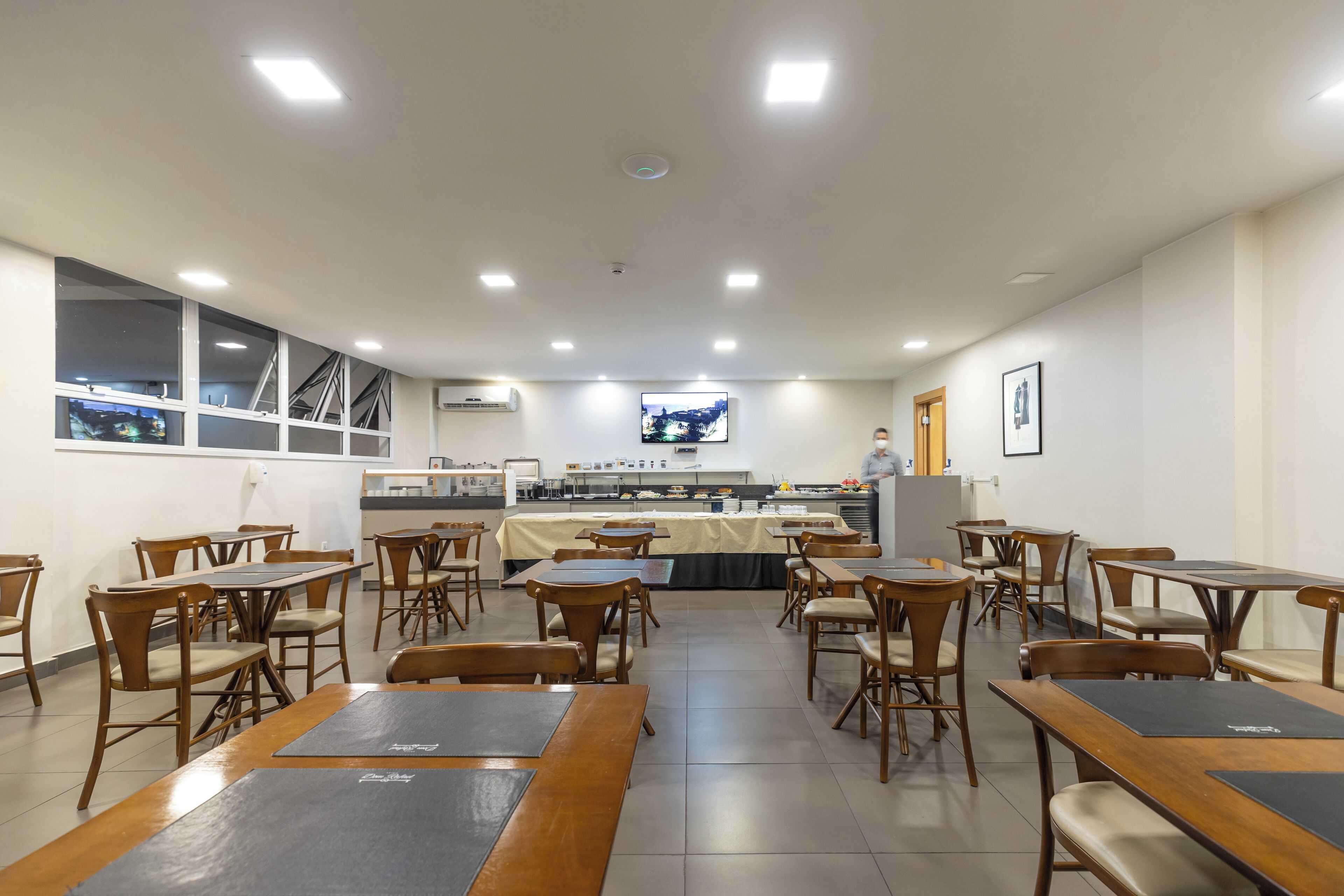 Un restaurante con mesas y sillas de madera, una zona de cocina y una pantalla de televisión en la pared.