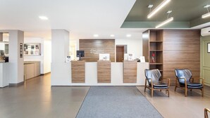 A modern reception area with a wooden reception desk, two leather chairs, and a grey carpet. - Hotel Dom Rafael Premium (Santa Maria)