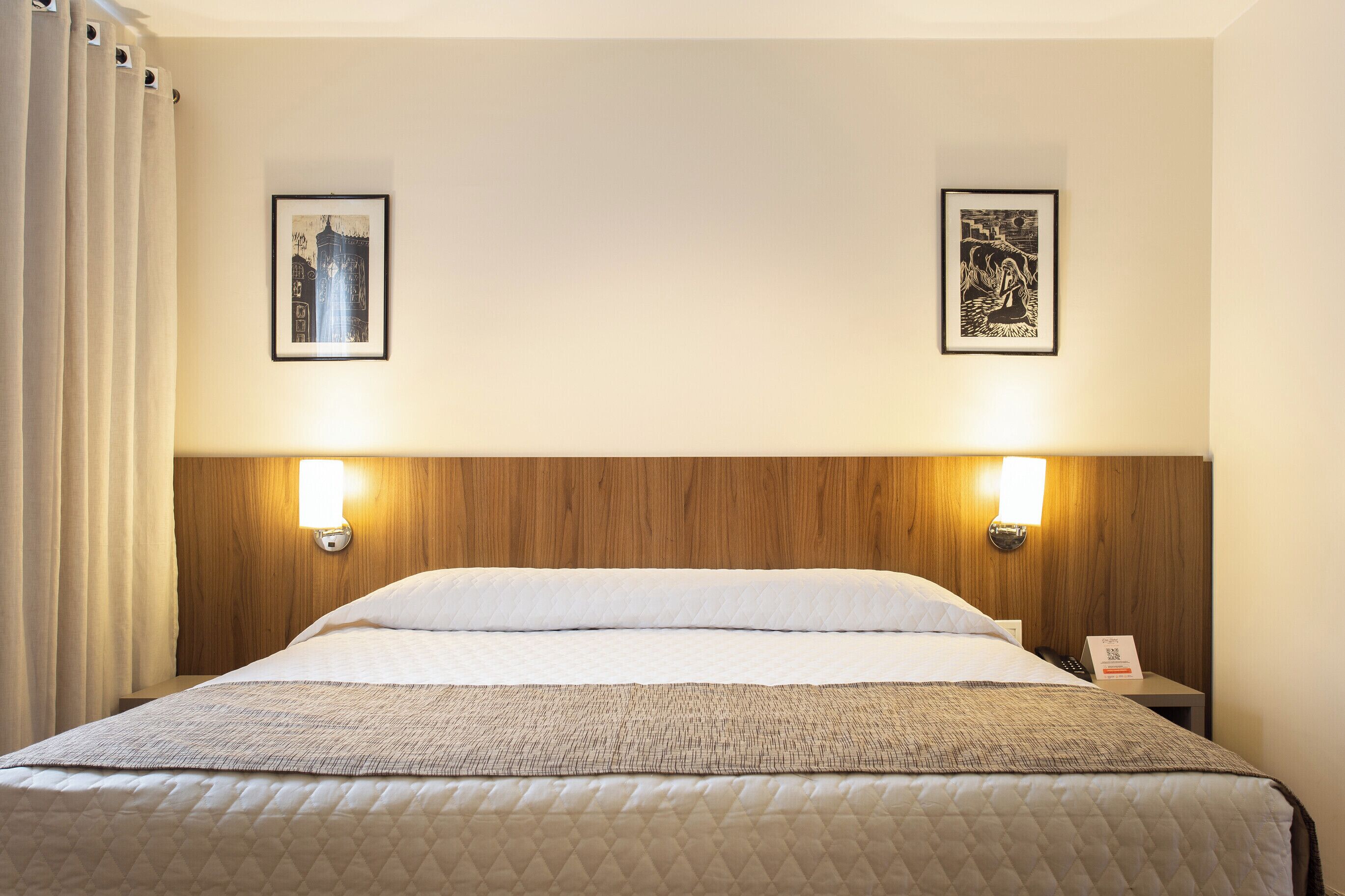 Habitación de hotel con cabecera de madera, dos lámparas de pared, cama con ropa blanca, manta acolchada y dos cuadros enmarcados.