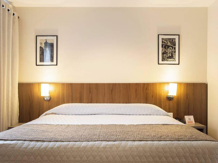 Een hotelkamer met een houten hoofdbord, twee wandlampen, een bed met wit beddengoed, een gewatteerde plaid en twee ingelijste kunstwerken aan de muur.