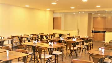 A classroom with wooden tables and chairs, a glass partition, and a wooden counter in the background.