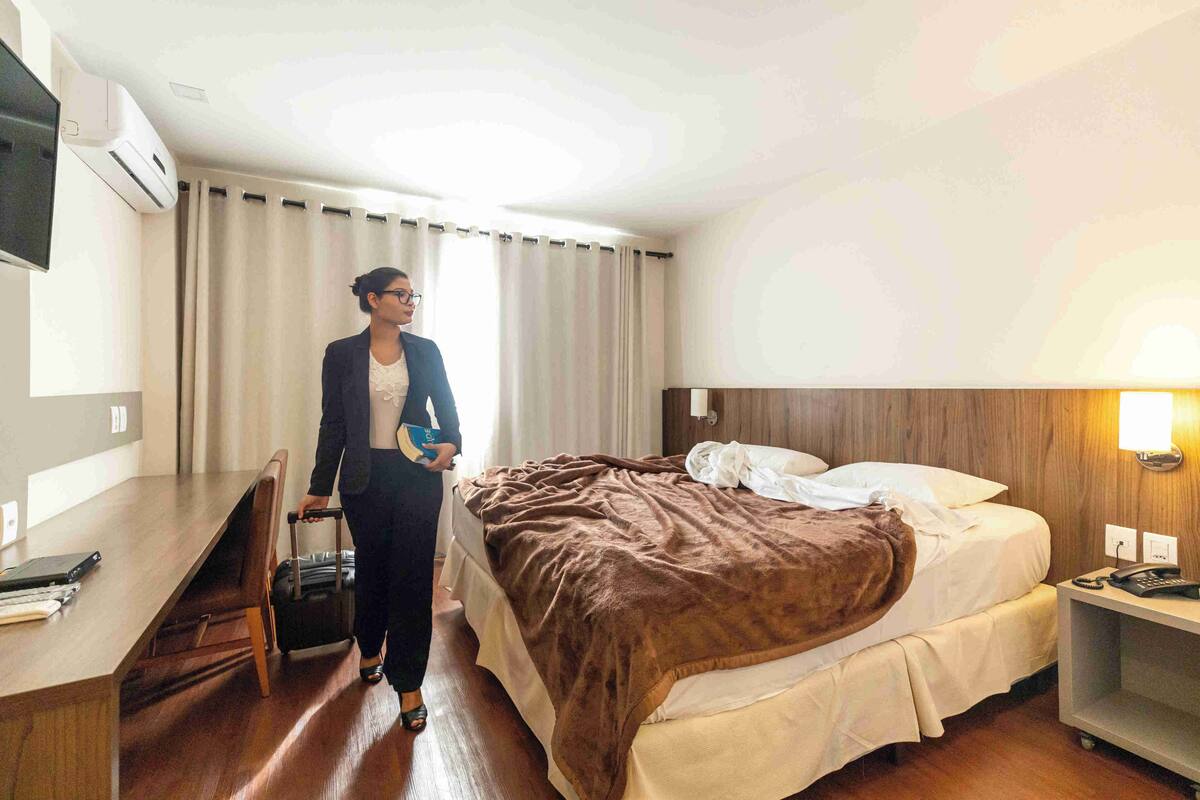 A hotel room with a bed, a desk, a chair, a suitcase, a TV, and a bedside lamp.