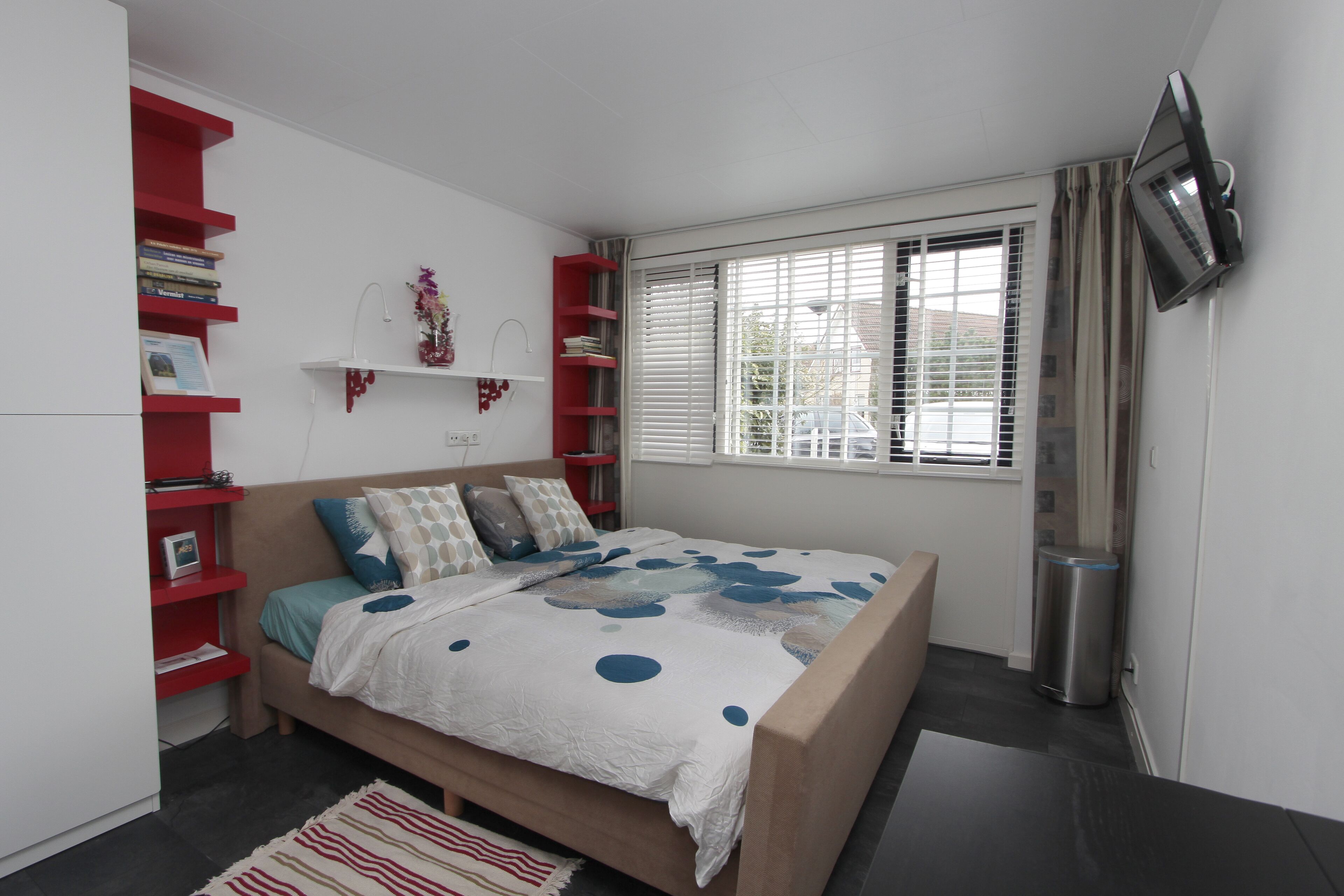 Un dormitorio con cama, una estantería roja, una ventana con persianas y un televisor montado en la pared.
