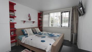 Un dormitorio con cama, una estantería roja, una ventana con persianas y un televisor montado en la pared.