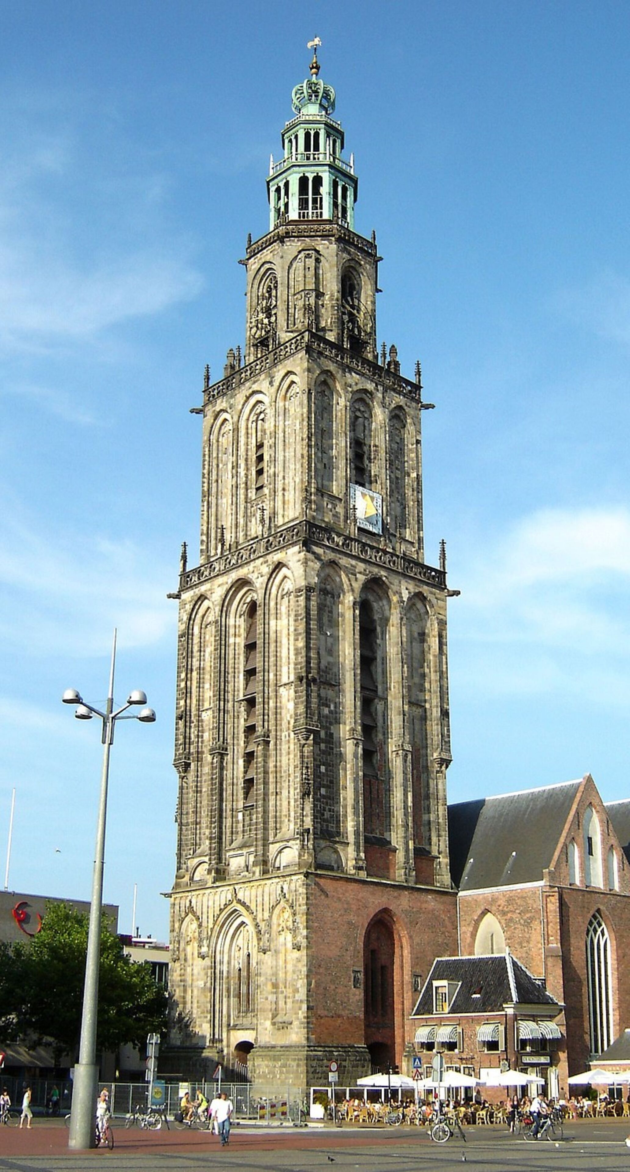 Ein historischer Turm mit einer Uhr, umgeben von einem Platz mit Außenbestuhlung und Fahrrädern.
