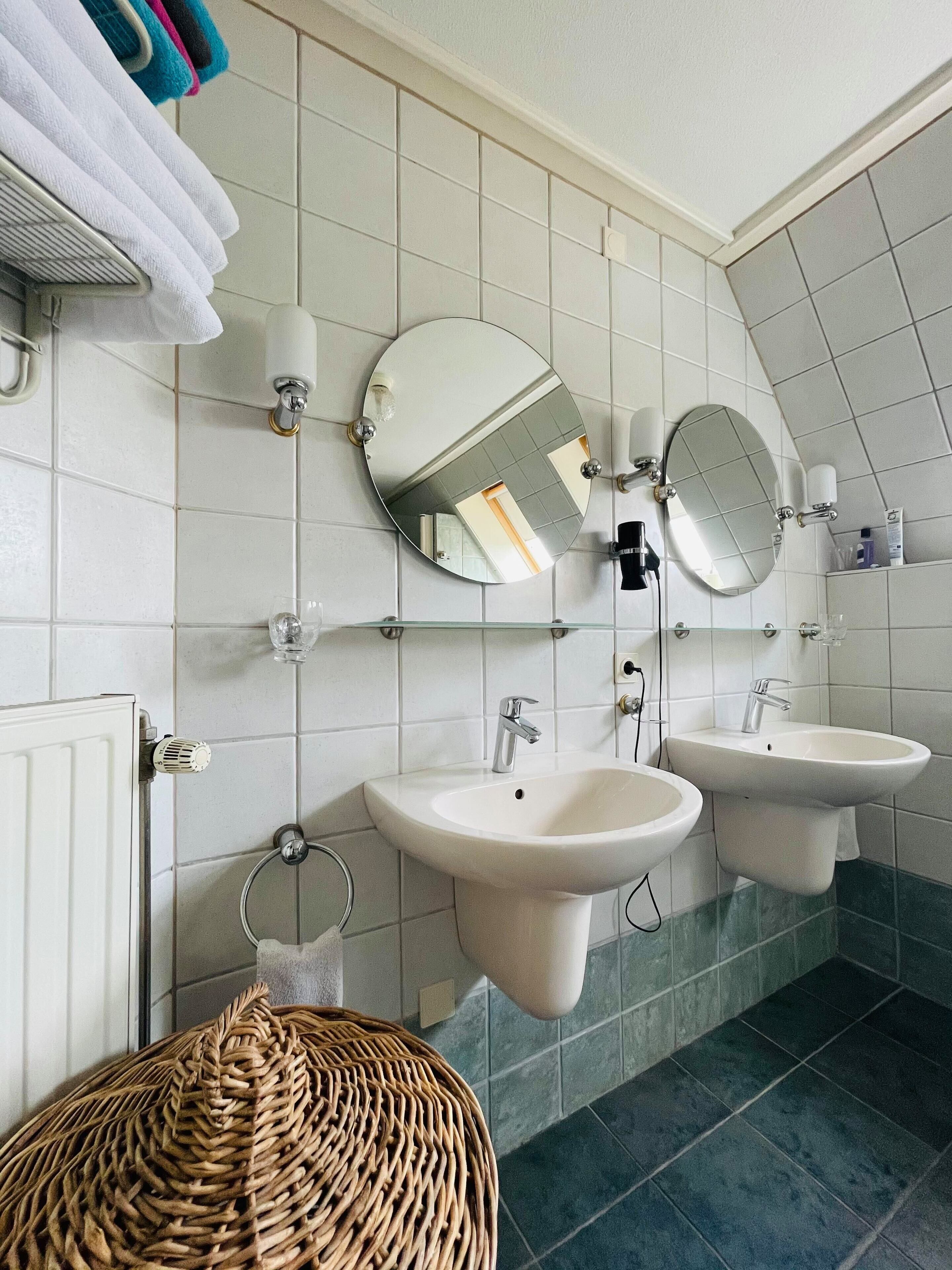Een badkamer met twee wastafels, twee spiegels, een handdoekrek met handdoeken en een rieten kruk.