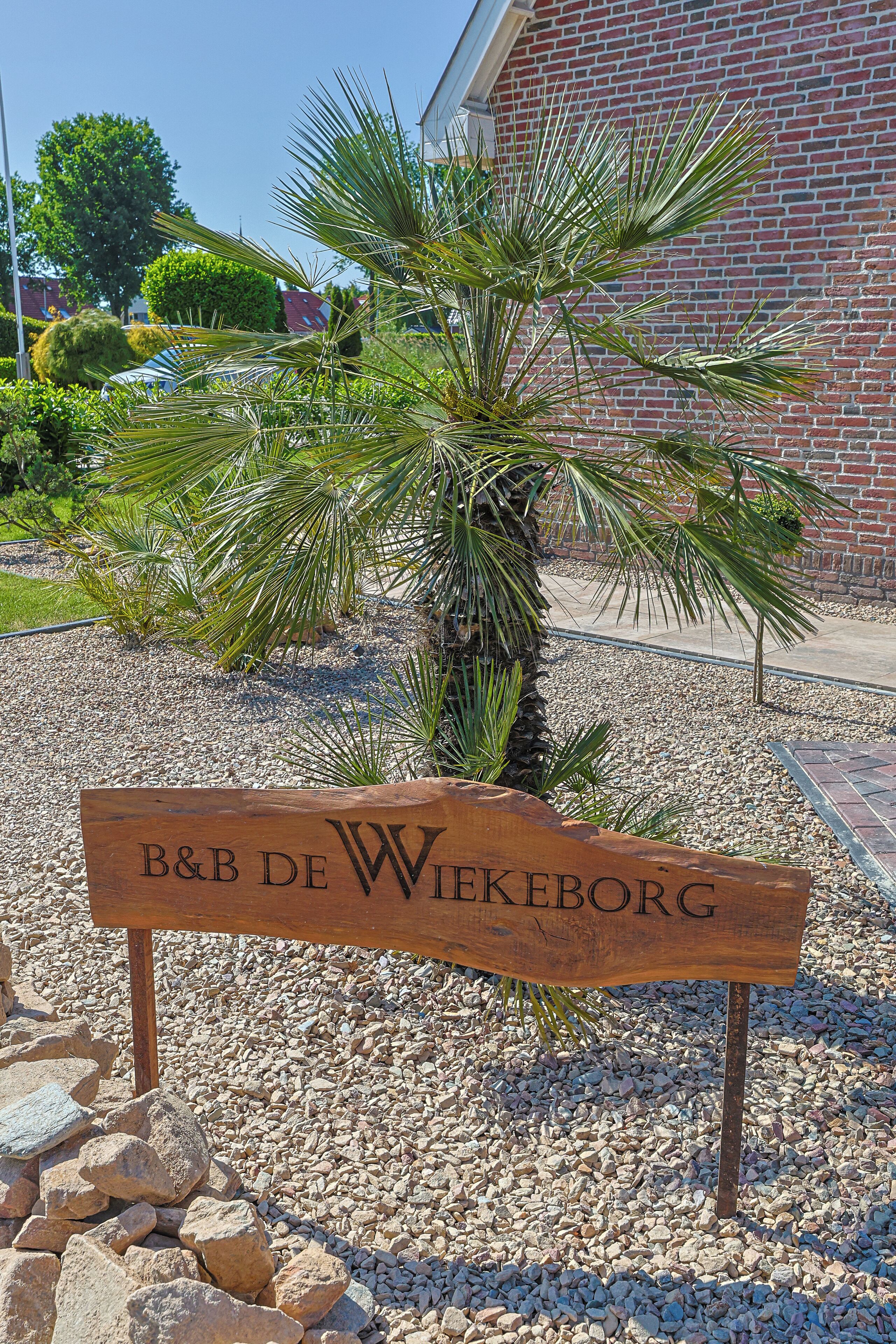 A wooden sign with "B&B DE WIEKEBORG" written on it, set against a backdrop of a brick building and a palm tree.