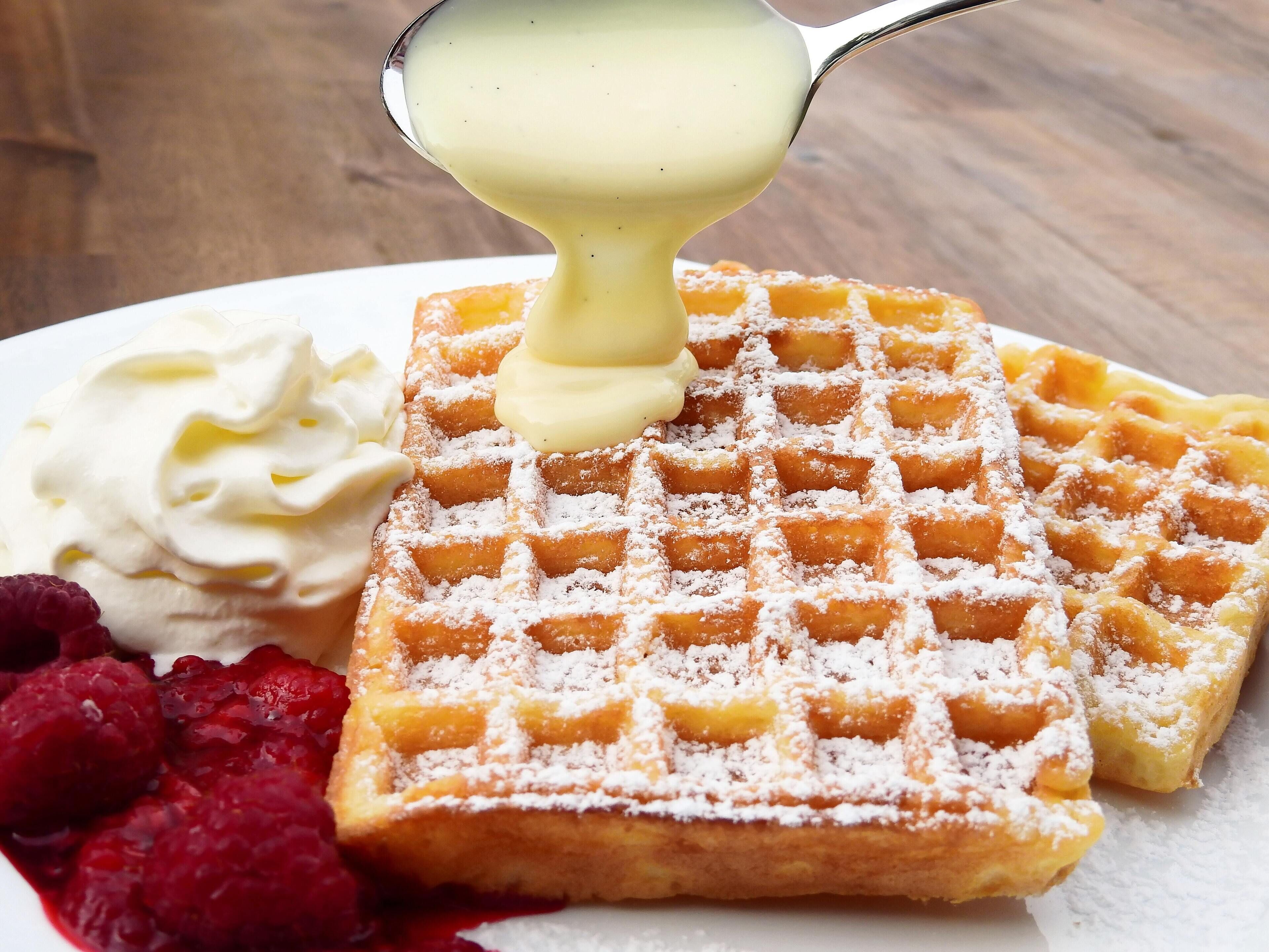 Une vue rapprochée d’un gaufre belge saupoudrée de sucre glace, surmontée d’une cuillerée de crème fouettée et de framboises fraîches, le tout présenté sur une assiette blanche.