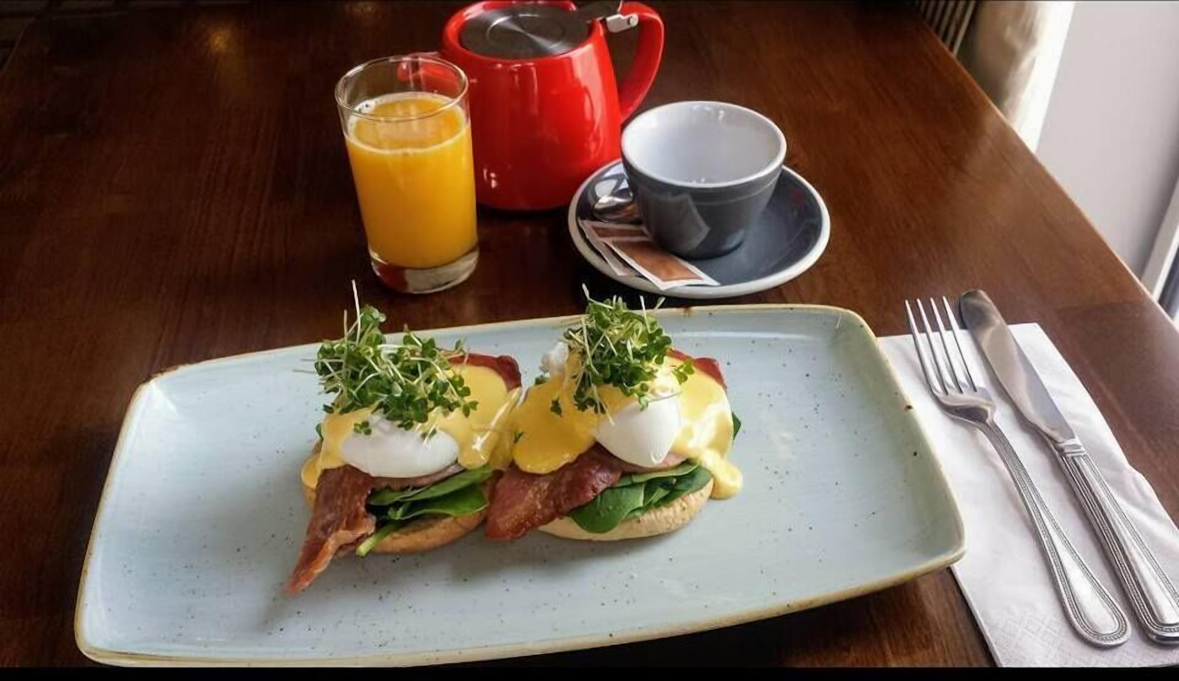 En frukosttallrik med ägg Benedict, en röd vattenkokare, ett glas apelsinjuice och en kopp på ett träbord.